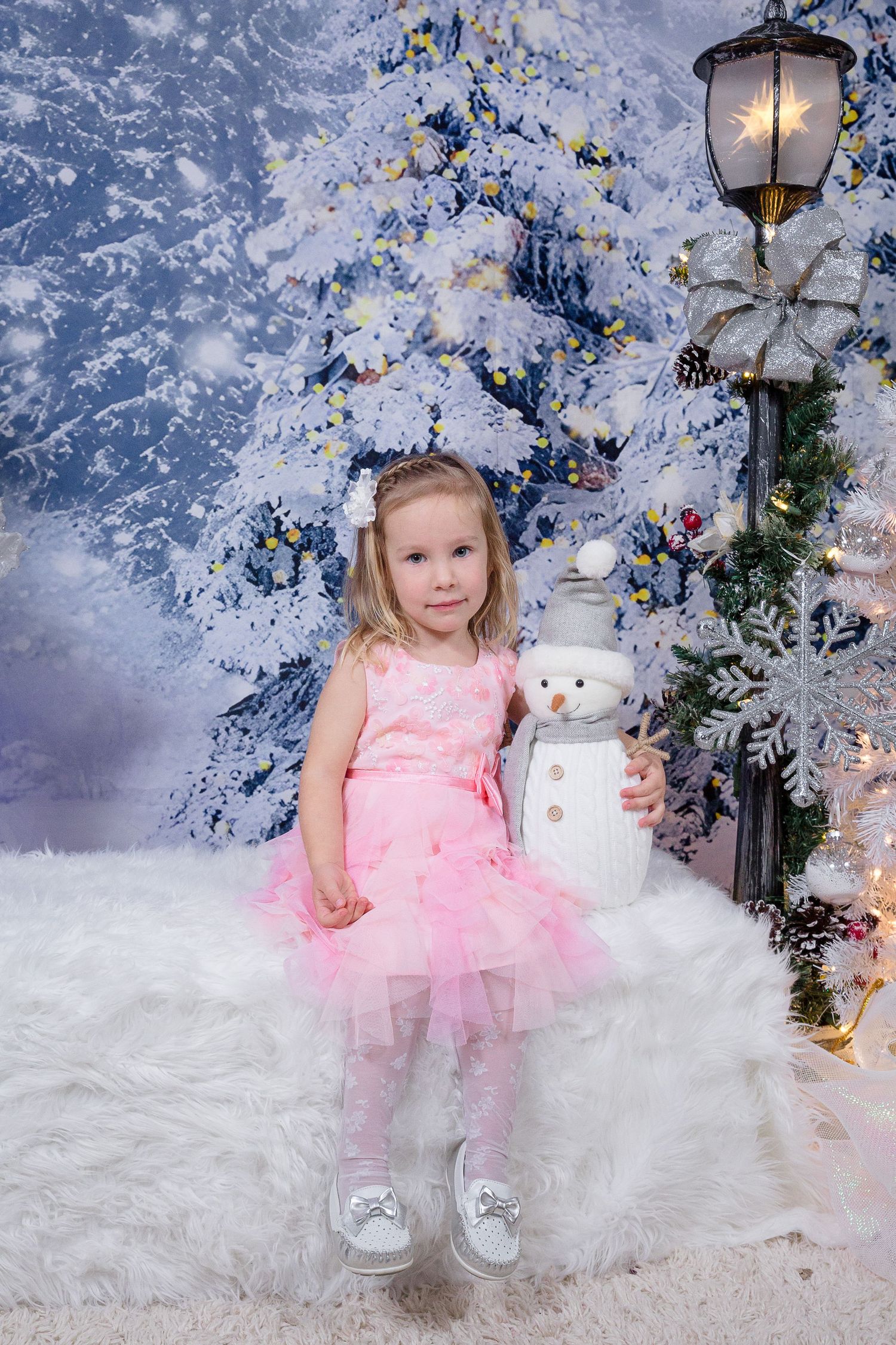 Child in pink tulle dress poses with snowman decoration against winter wonderland backdrop.
