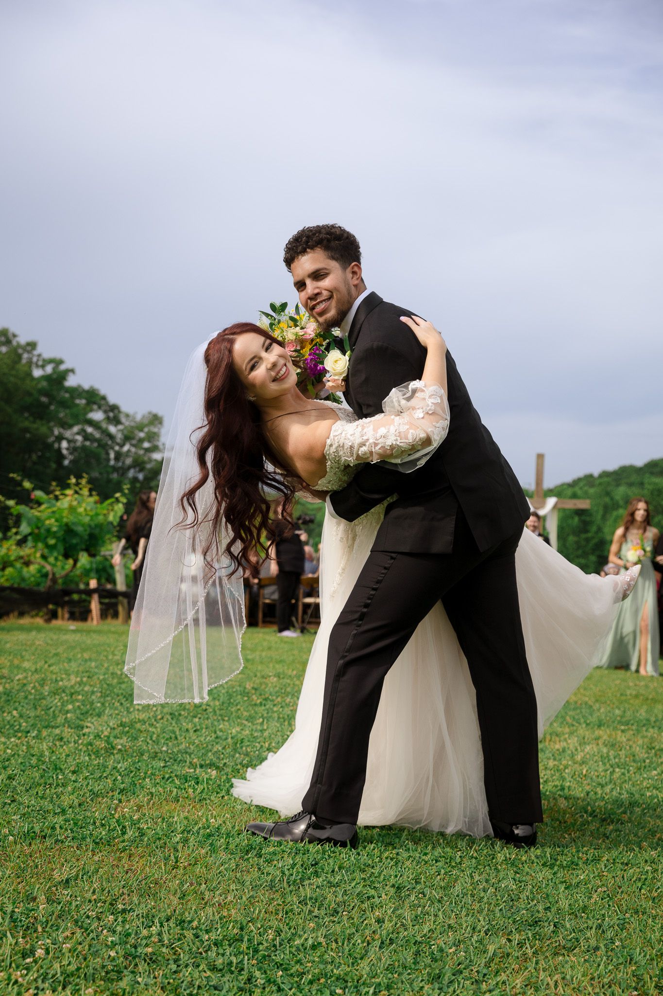 Joyful bride and groom dancing outdoors after wedding ceremony, bride in white gown with veil holding colorful bouquet.