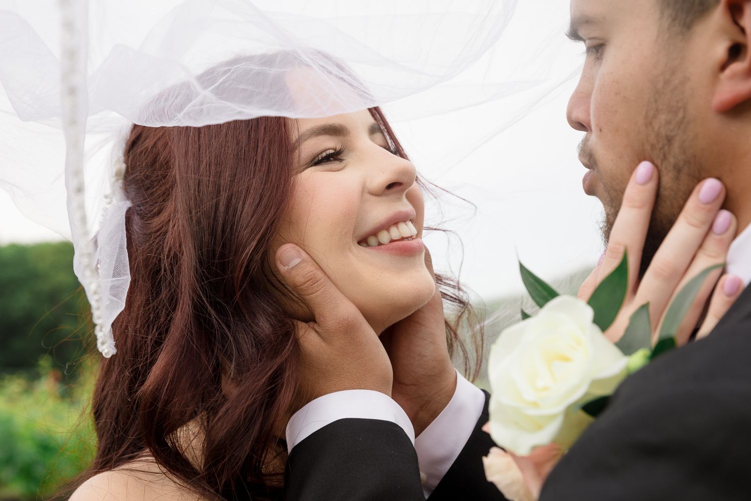 A smiling bride in a white veil gazes up at her groom during a romantic wedding photo shoot outdoors.