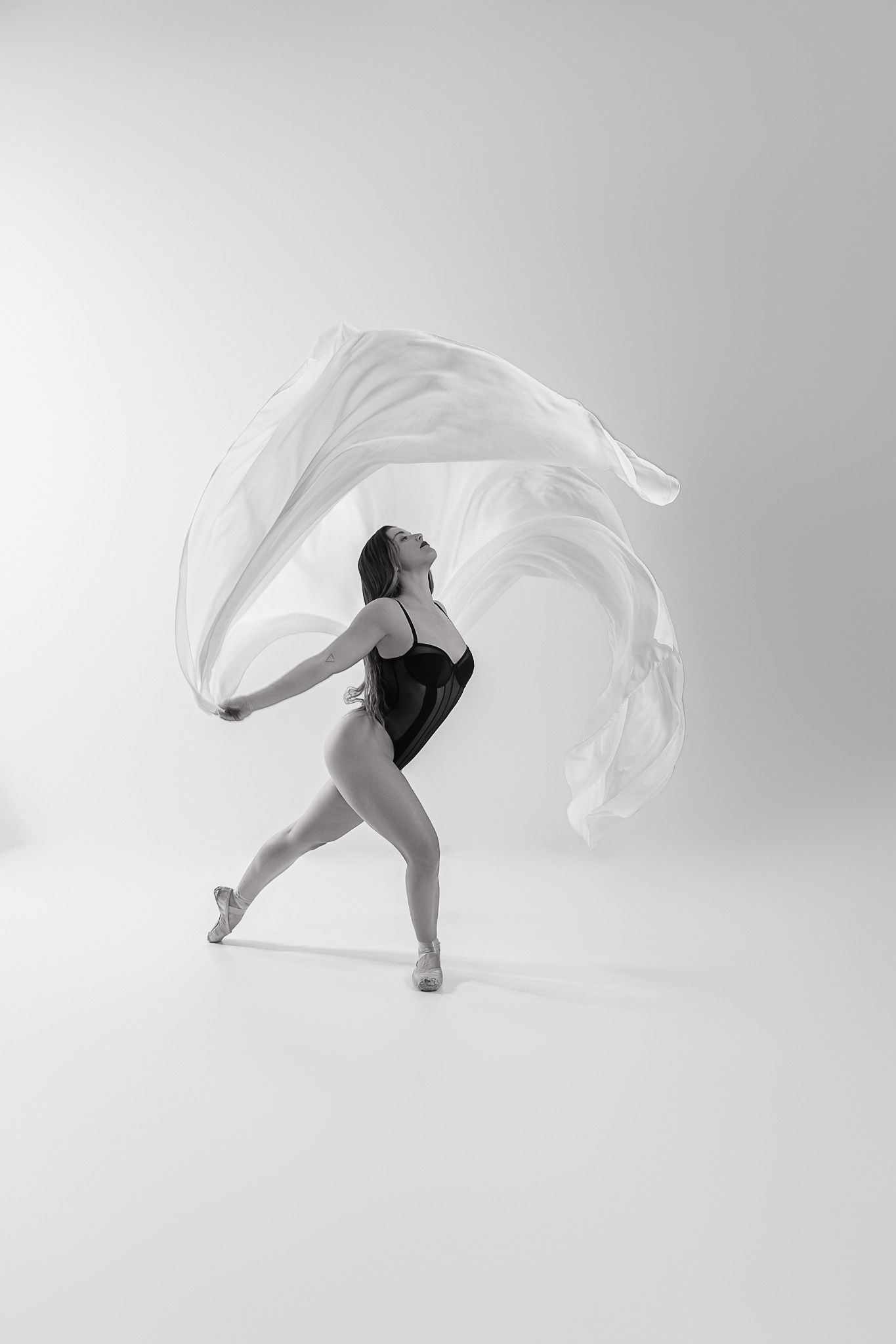 A dancer in black leotard performs graceful movements with flowing white fabric against a light background.