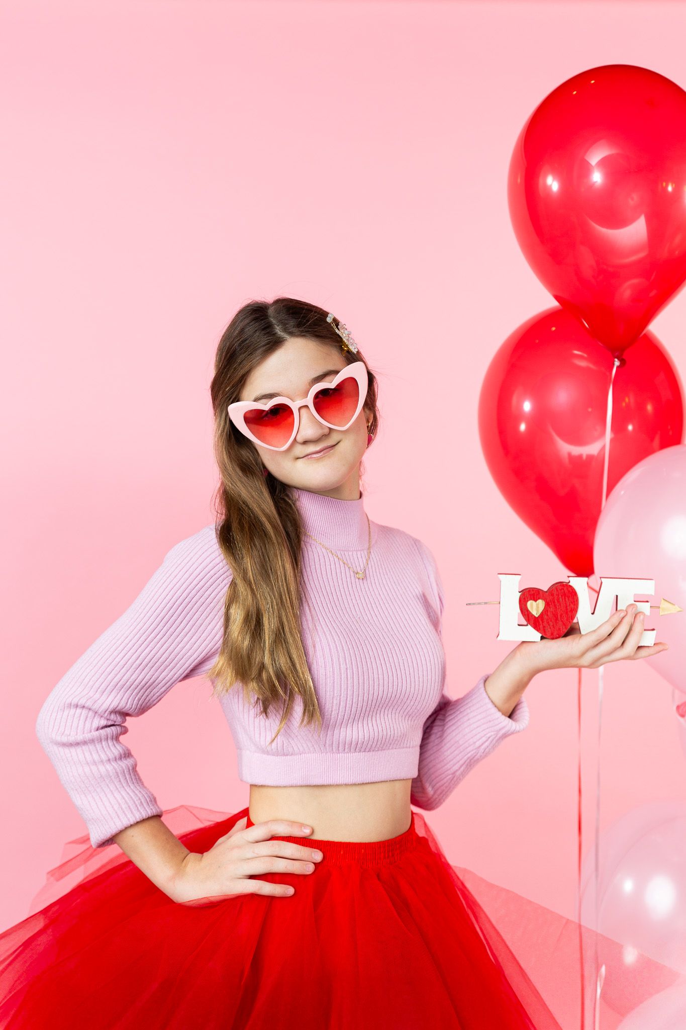 Person in pink sweater and red tulle skirt posing with heart sunglasses and Valentine balloons.