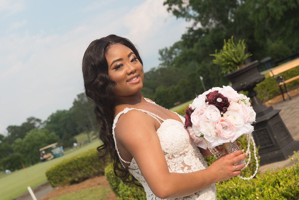 Smiling bride in lace wedding dress holding pink and burgundy bouquet outdoors at a golf course venue.