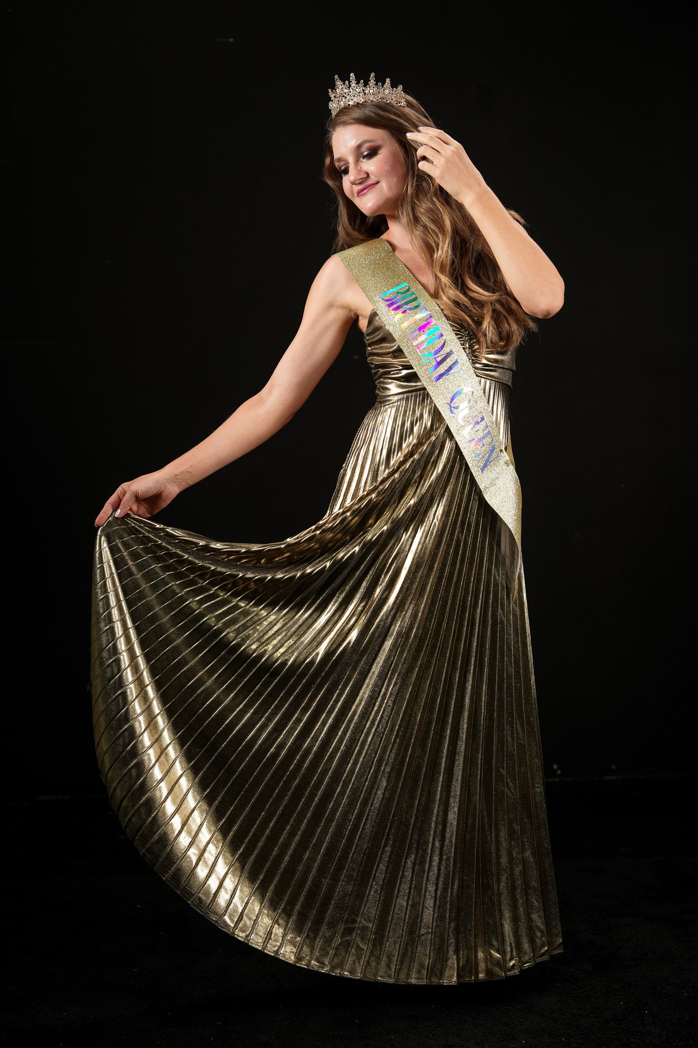 Young woman in gold pleated gown wearing pageant crown and sash poses gracefully against black background.