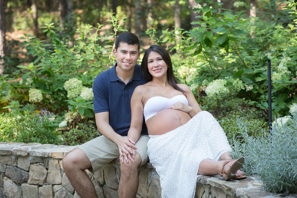 Outdoor maternity session featuring couple relaxing on stone wall with garden backdrop.
