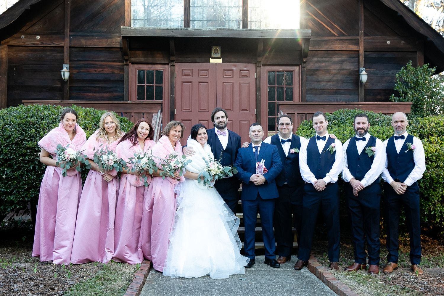 Wedding party posing outside rustic wooden chapel, bridesmaids in pink gowns, groomsmen in navy suits, bride and groom centered.