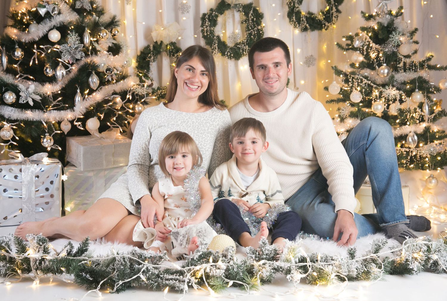 Family holiday photoshoot scene with Christmas trees and twinkling lights in the background.