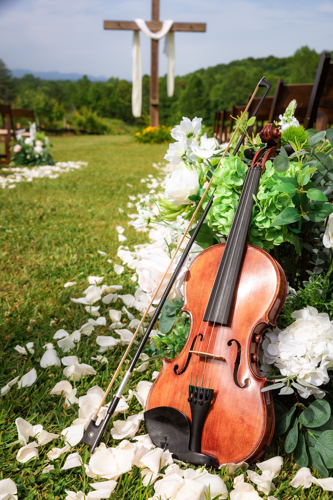 Violin and bow resting against white floral arrangement on grass with wooden cross and green hills in background.