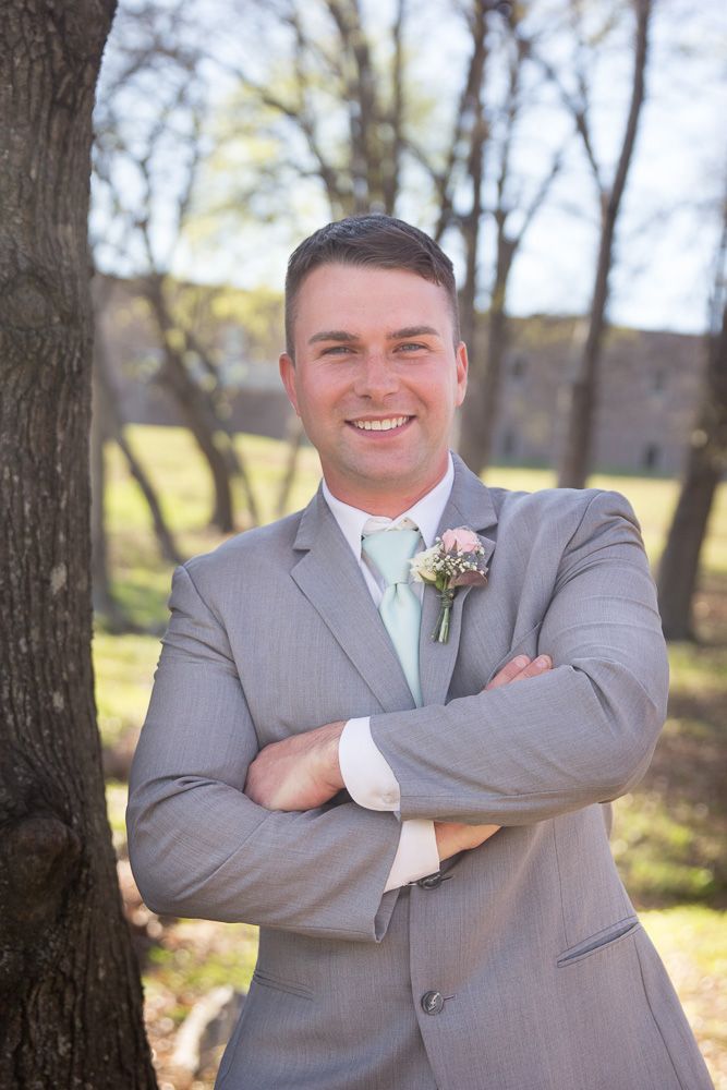 Smiling groom in gray suit with mint tie and floral boutonniere, arms crossed, posing outdoors near trees.