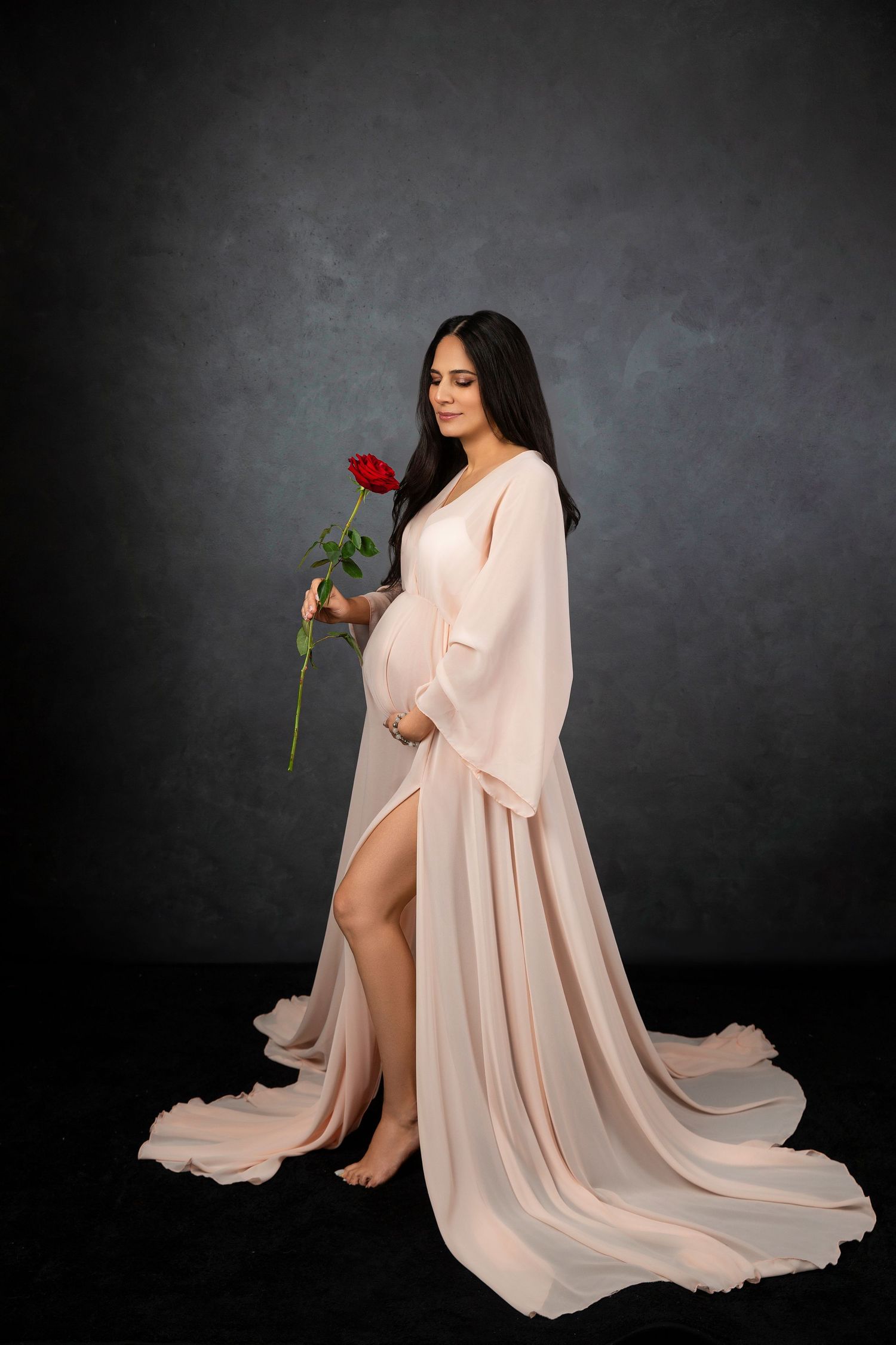 Graceful maternity pose with a long blush-colored gown and red rose on dark backdrop.