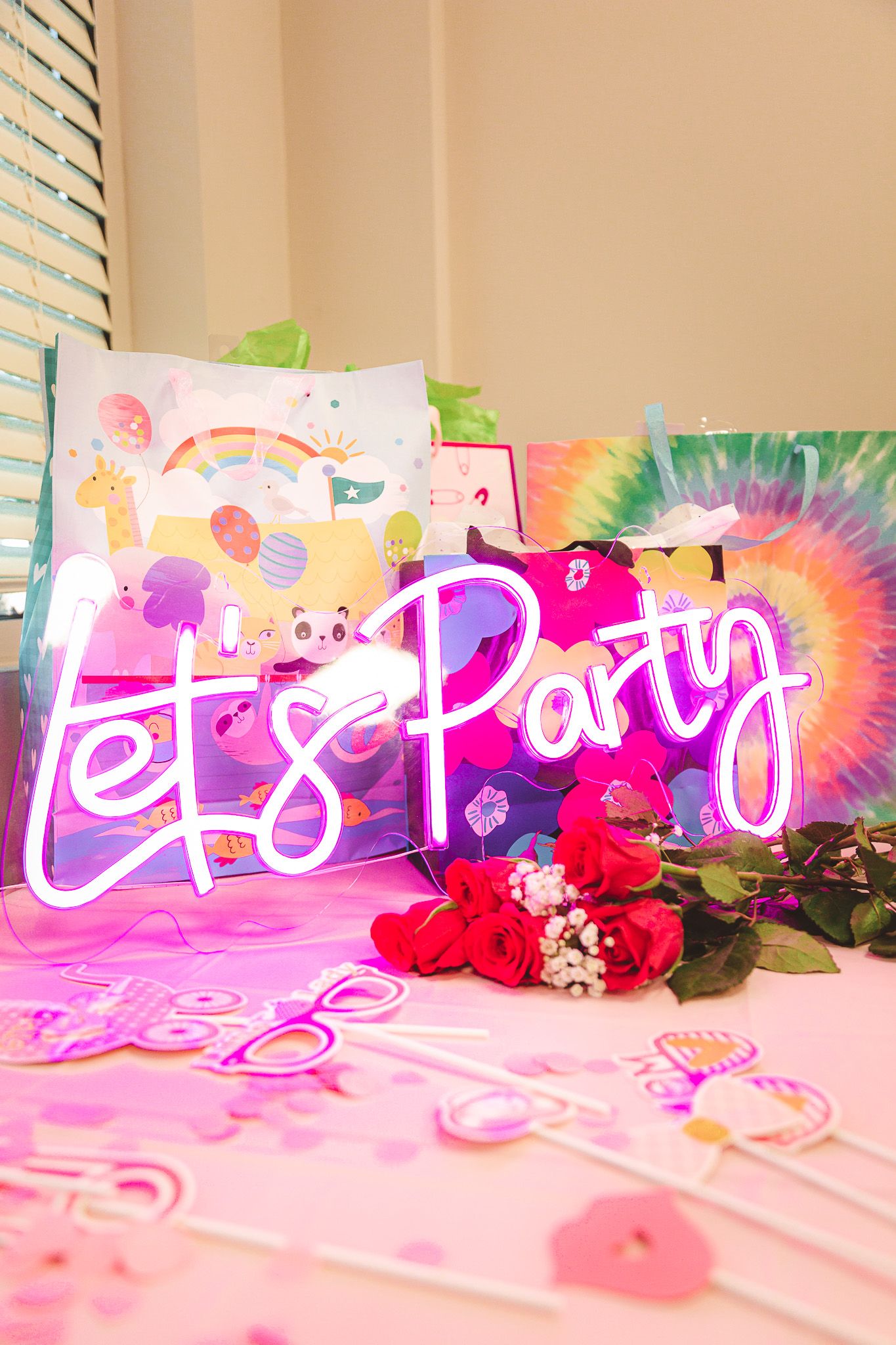 Pink neon 'Let's Party' sign displayed with roses and tie-dye decorations against a beige wall.