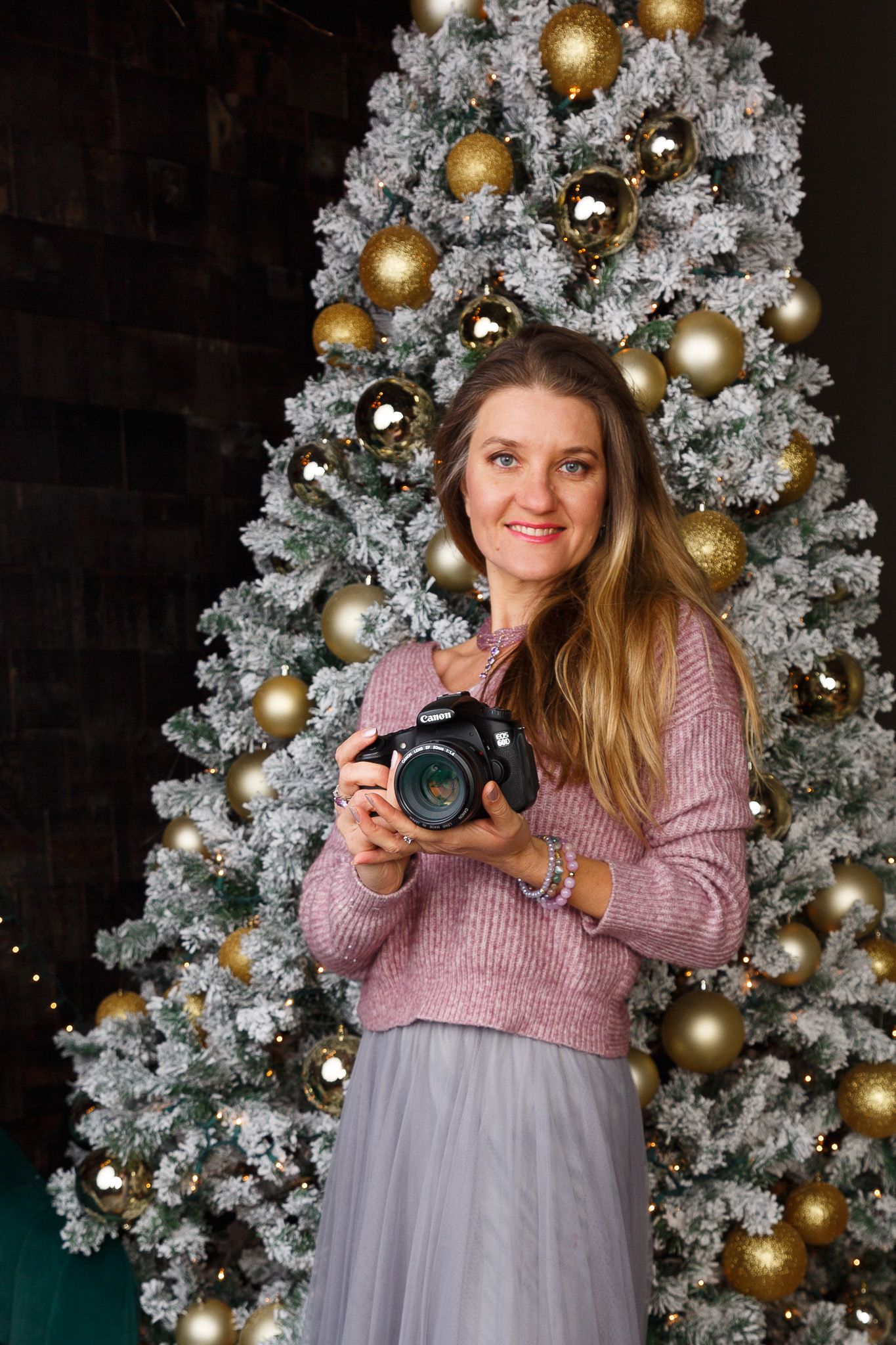 A photographer in a pink sweater holds a camera while standing in front of a flocked Christmas tree with gold ornaments.