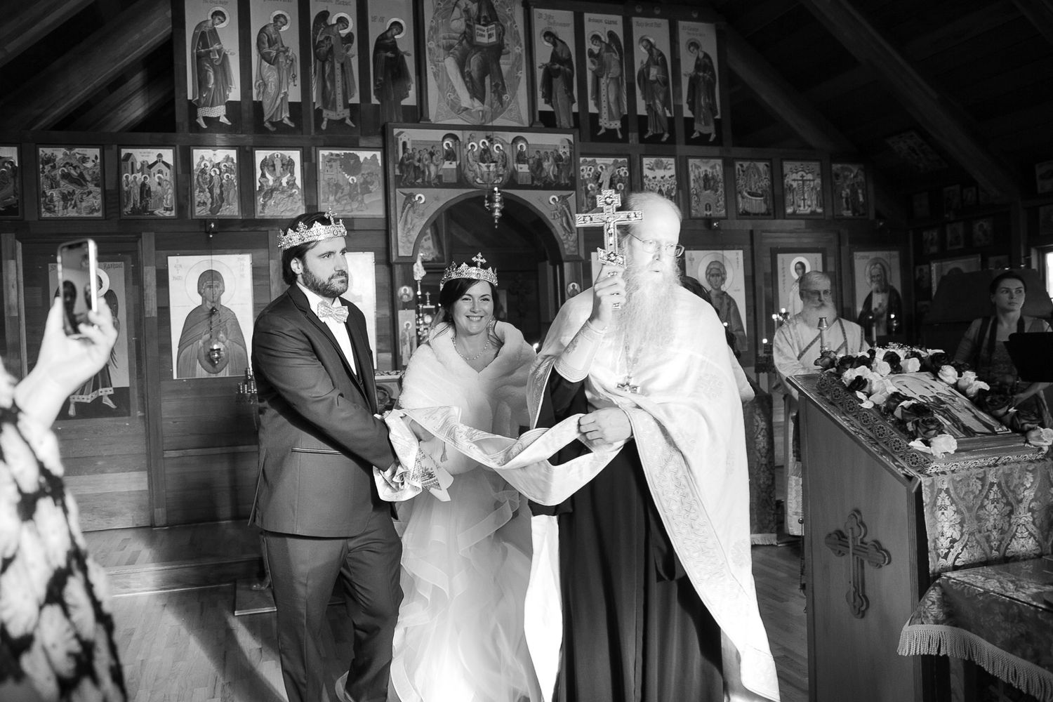 Black and white Orthodox wedding ceremony with couple wearing crowns and priest reading from scripture in decorated church.