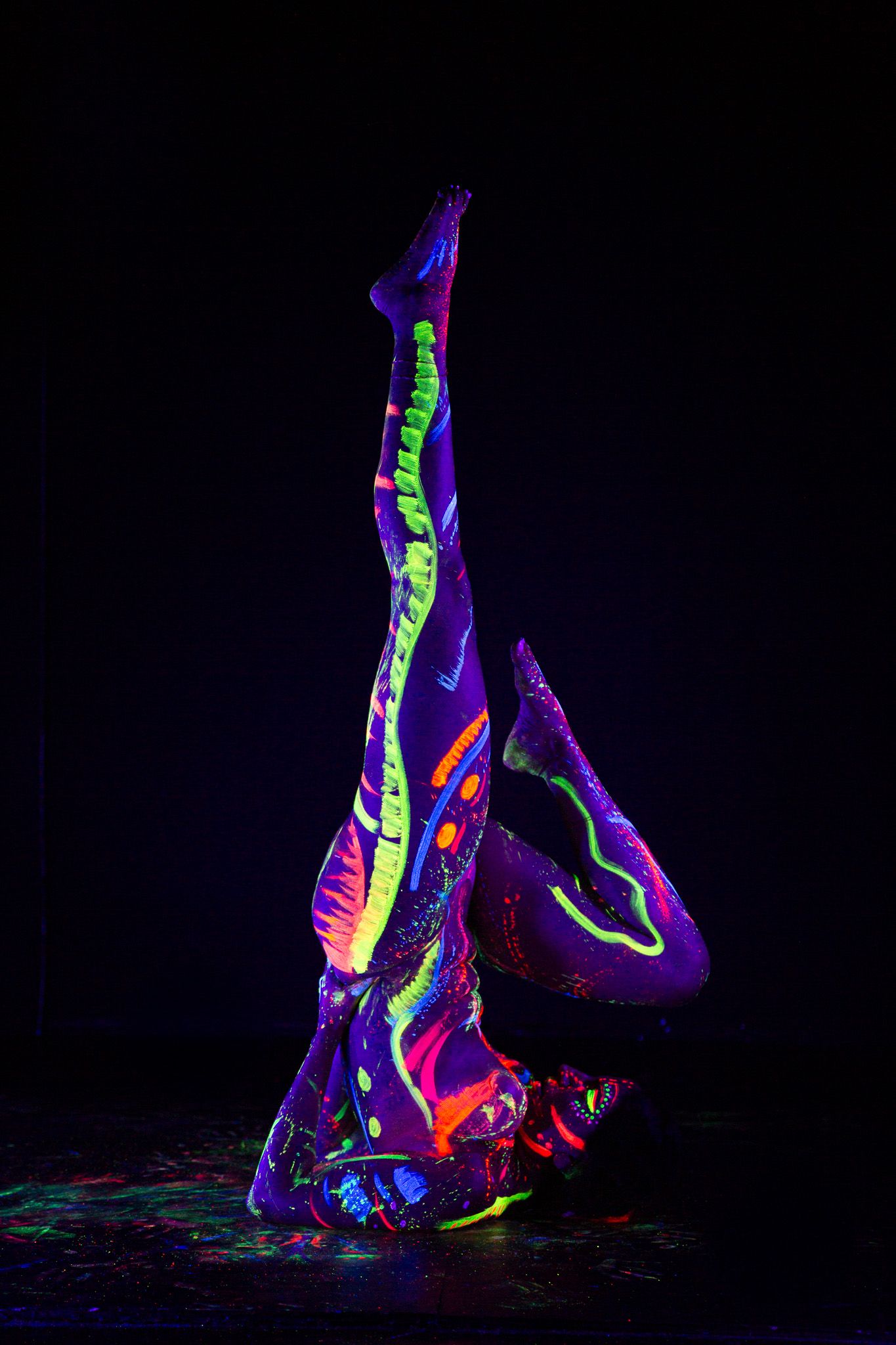 Acrobatic figure painted in fluorescent colors performing inverted pose in dark setting.