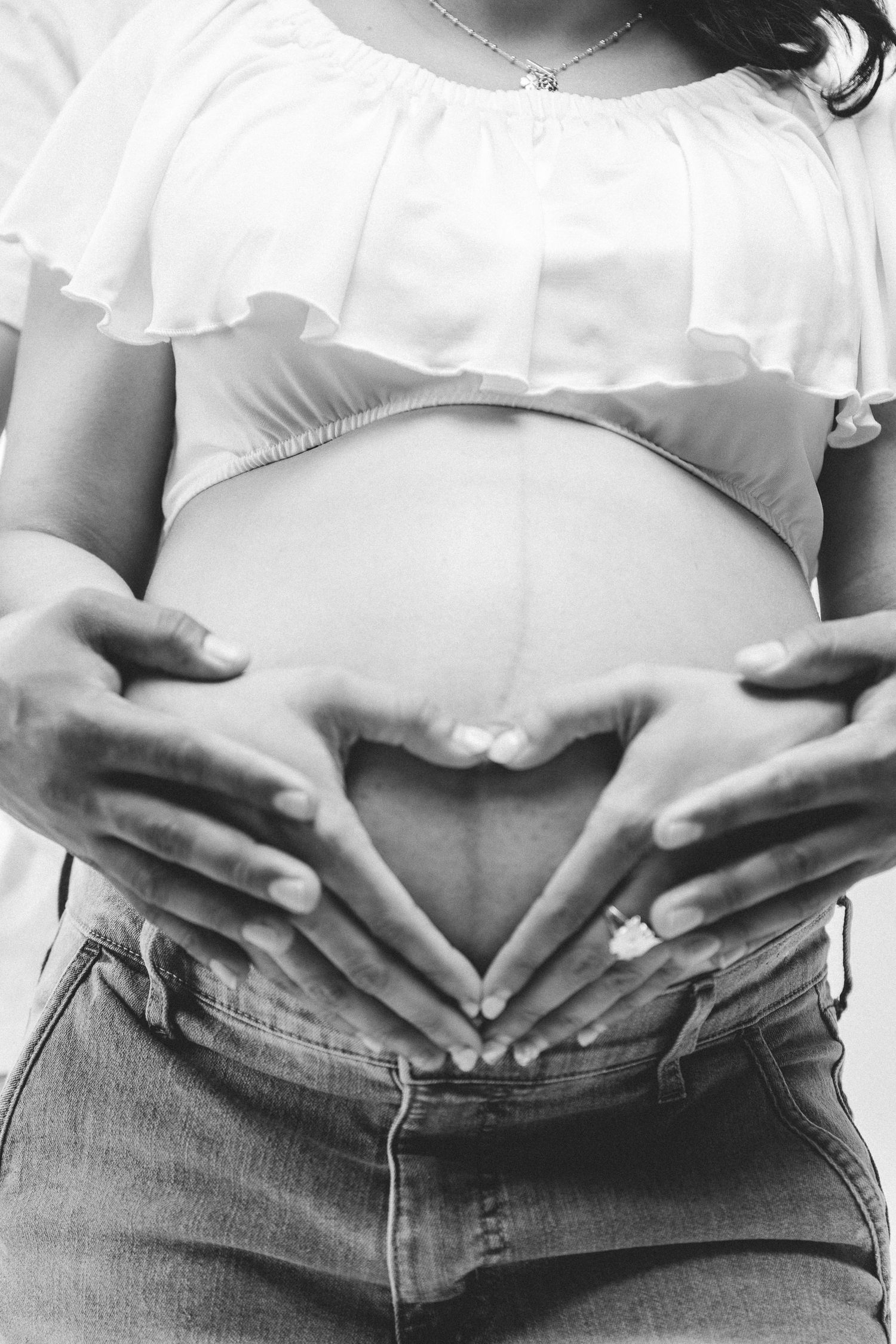 Black and white photo of pregnant woman forming heart shape with hands on baby bump wearing white top and jeans.