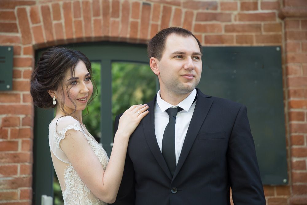 Elegant wedding couple posing outdoors, bride in lace dress touching groom's shoulder in black suit and tie.