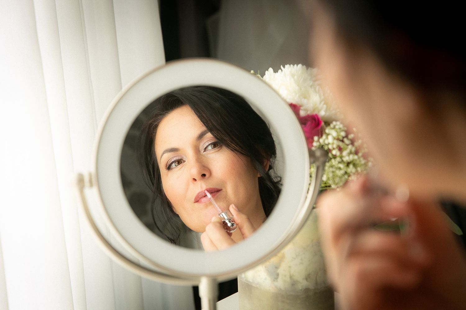 Bride applying lipstick while looking into a round vanity mirror during wedding day preparation.
