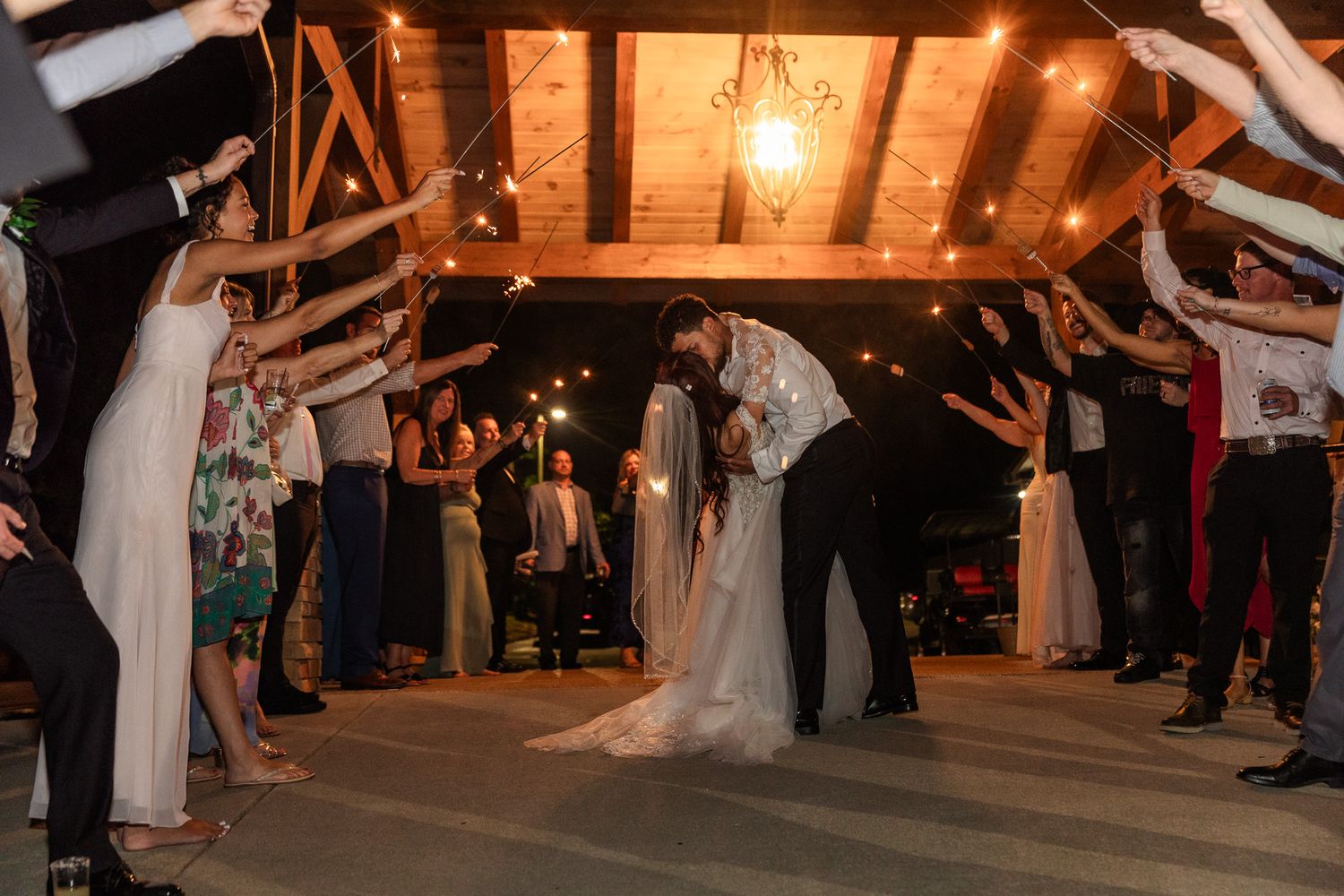 Wedding couple shares a kiss under sparklers held by guests in a warmly lit rustic barn venue.