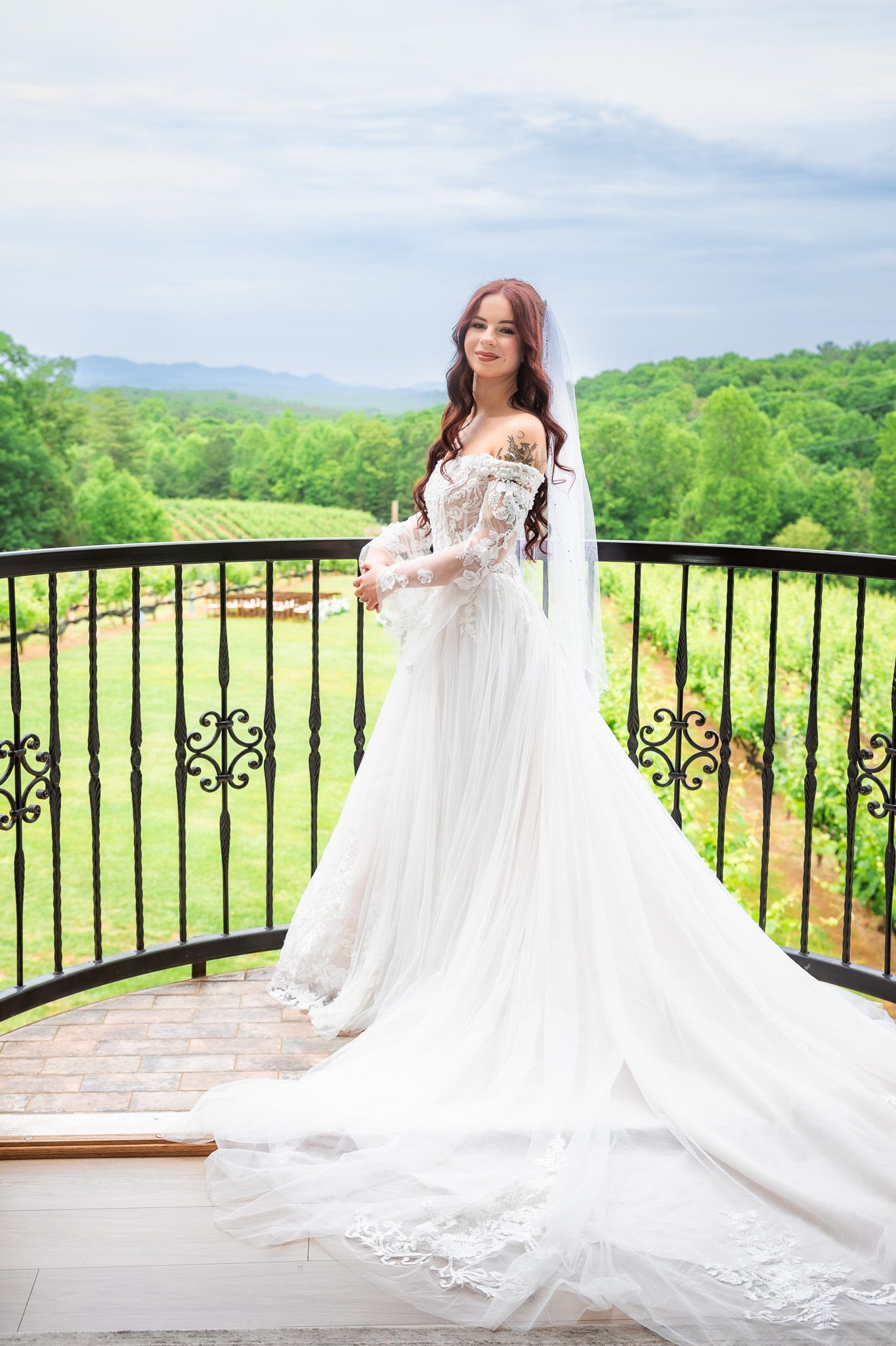 Bride in flowing lace wedding dress with veil on balcony overlooking vineyard.