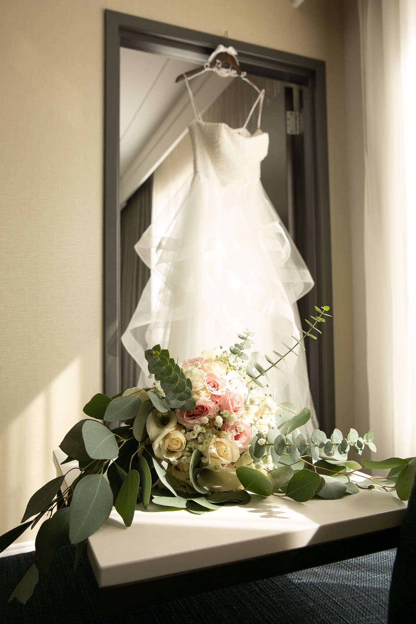 White wedding dress hanging on door frame with bridal bouquet of pink and cream roses with eucalyptus on table.