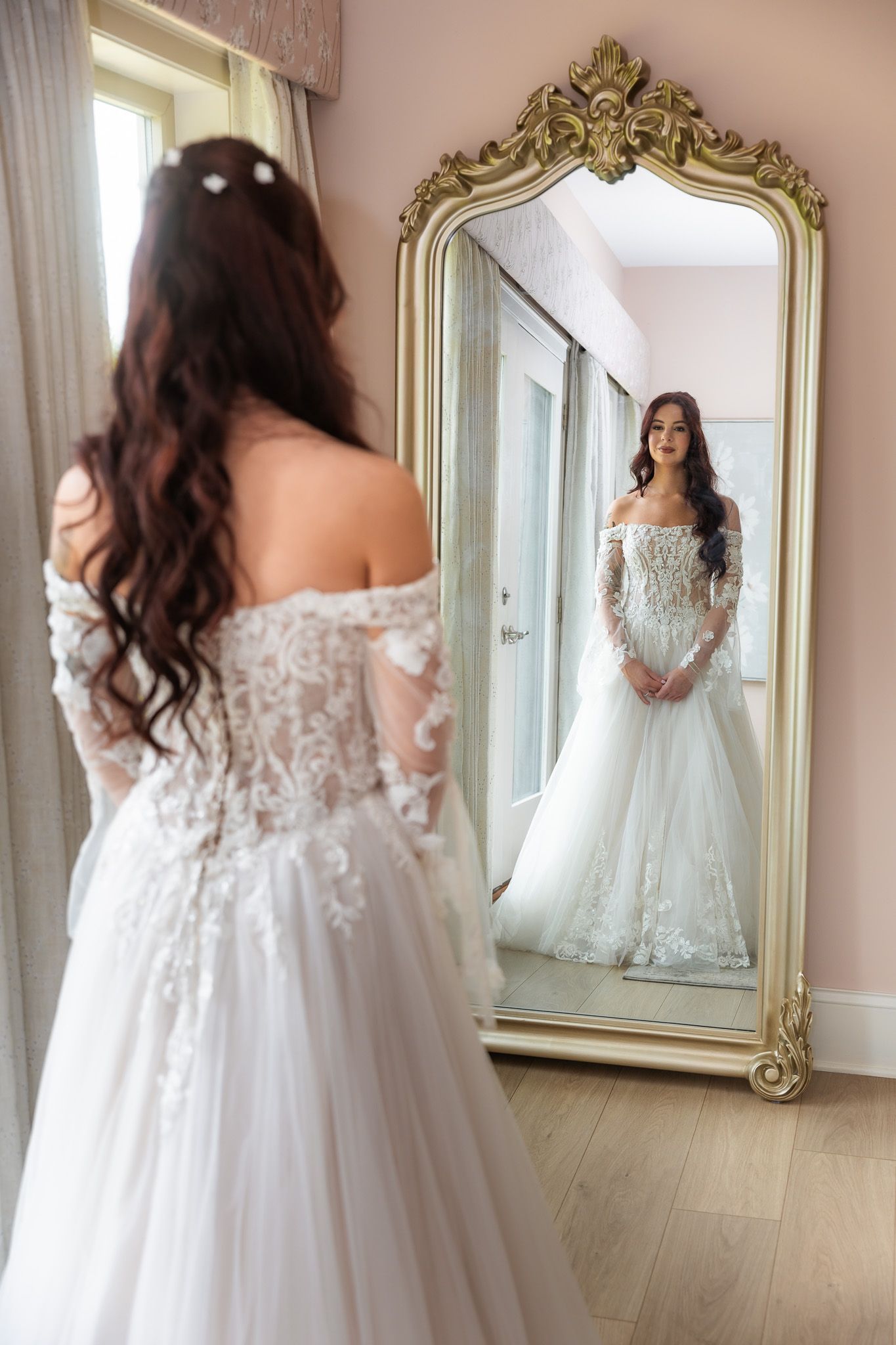 Bride in off-shoulder lace gown viewing reflection in ornate gold mirror.