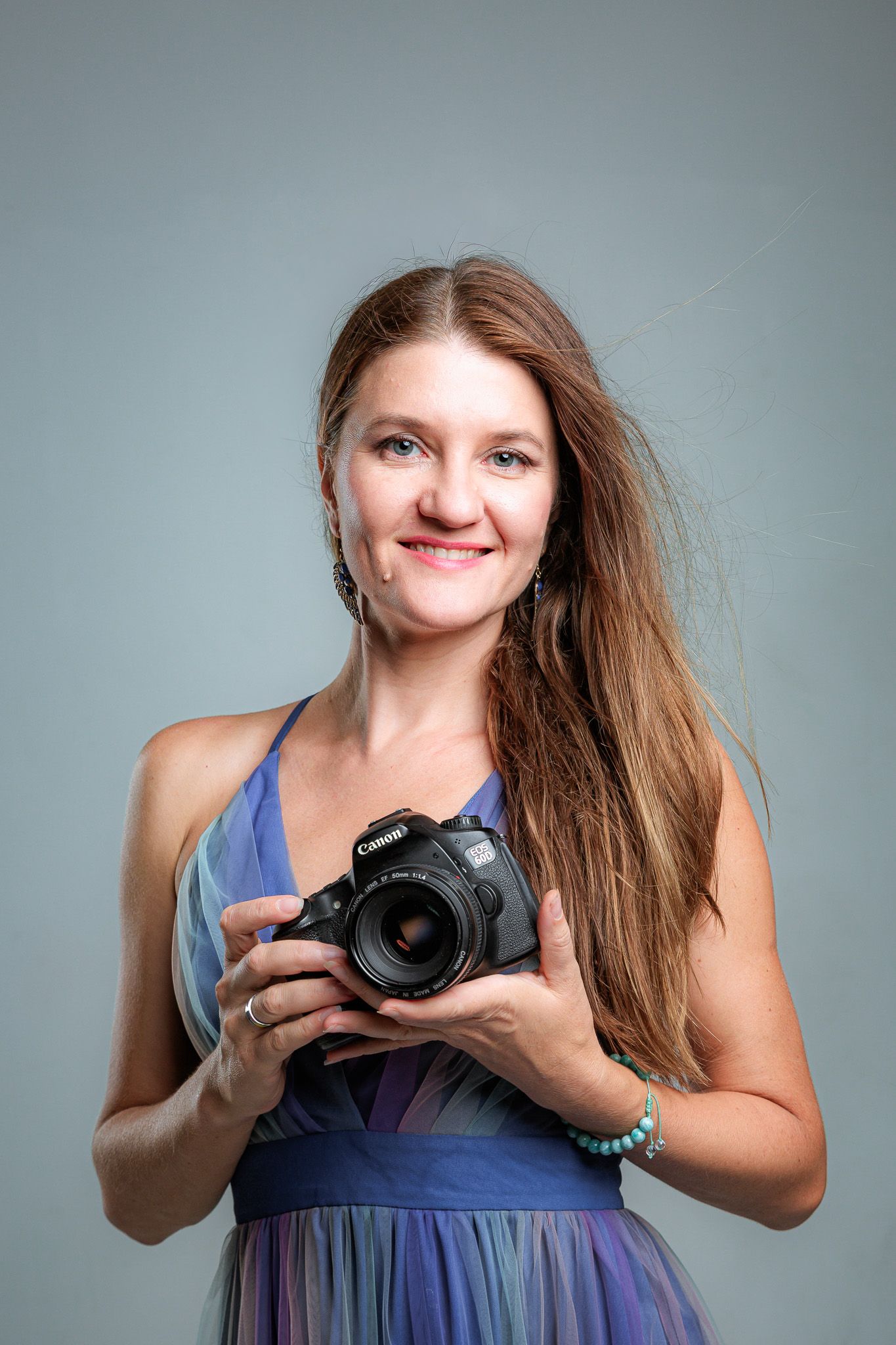 Photographer in a purple dress holds a DSLR camera while smiling against gray backdrop.