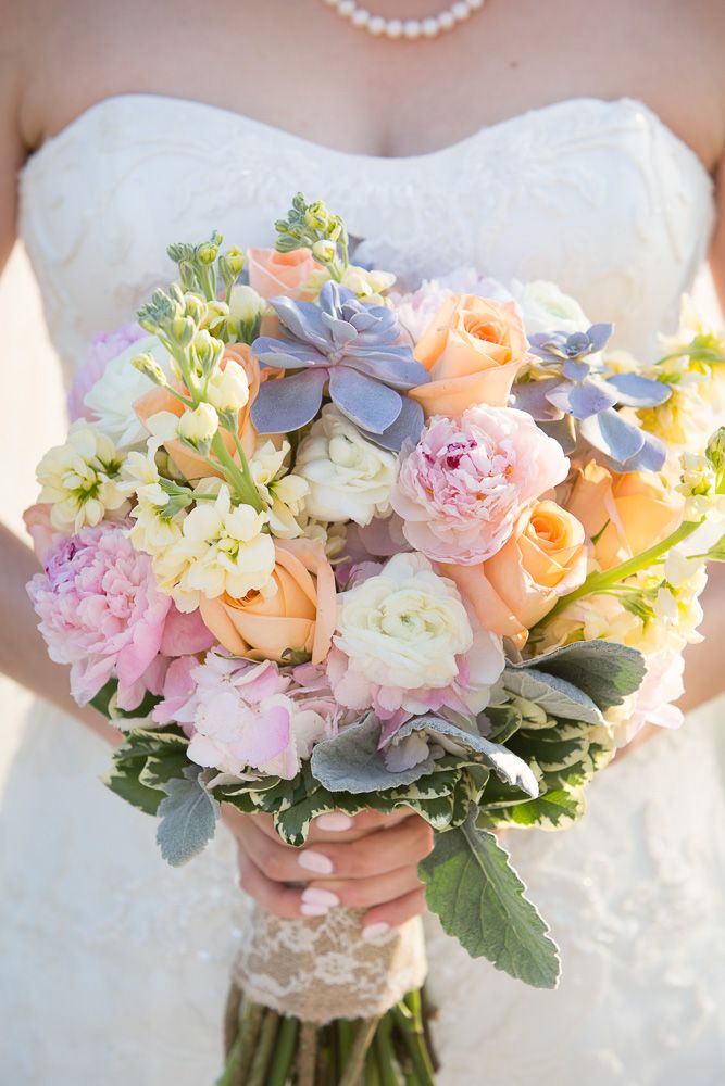 Bride holding a pastel wedding bouquet with peach roses, pink peonies, succulents, and white blooms wrapped in lace ribbon.