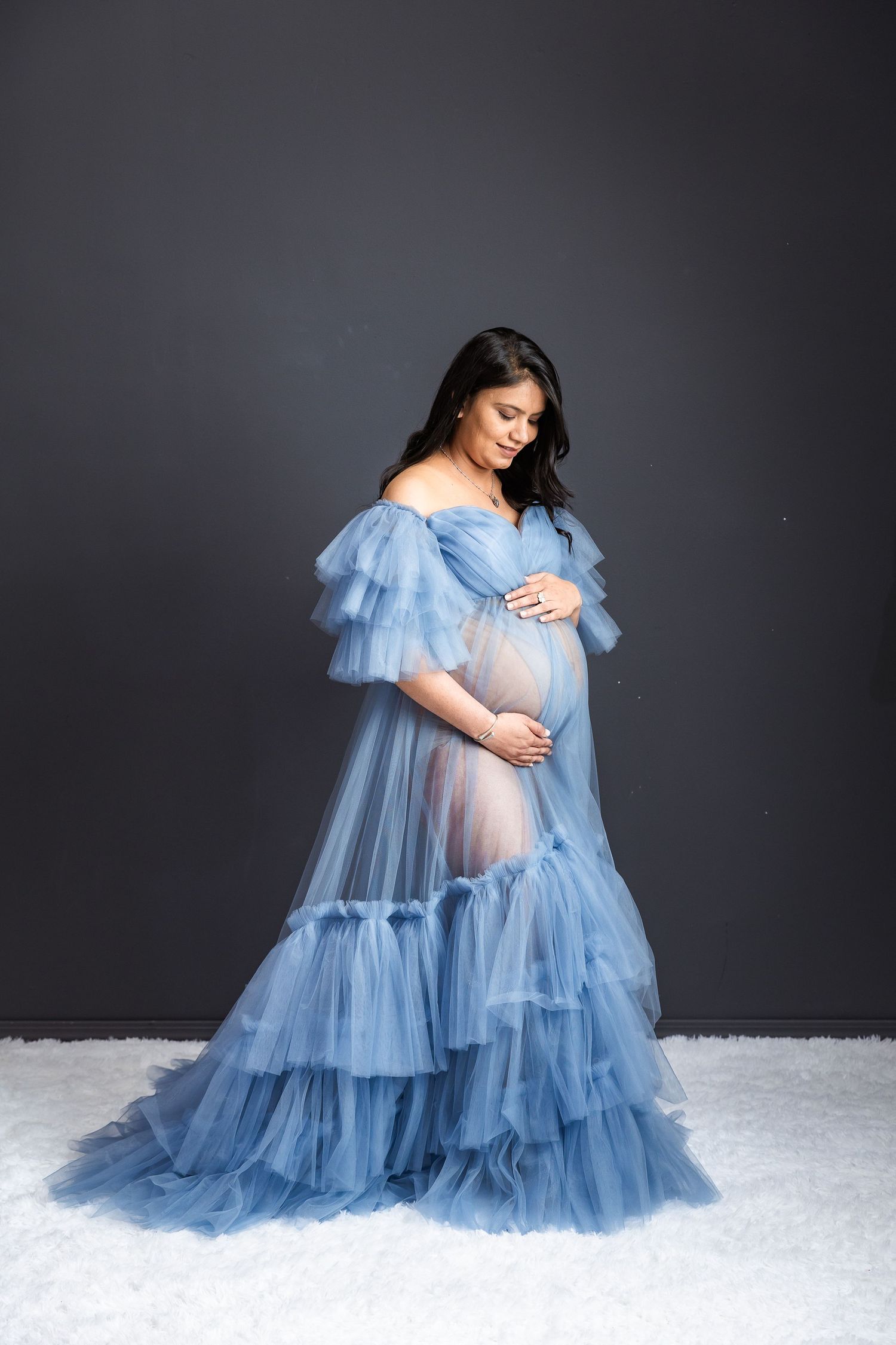 Pregnant woman in elegant blue tulle off-shoulder gown cradling baby bump against gray studio backdrop.