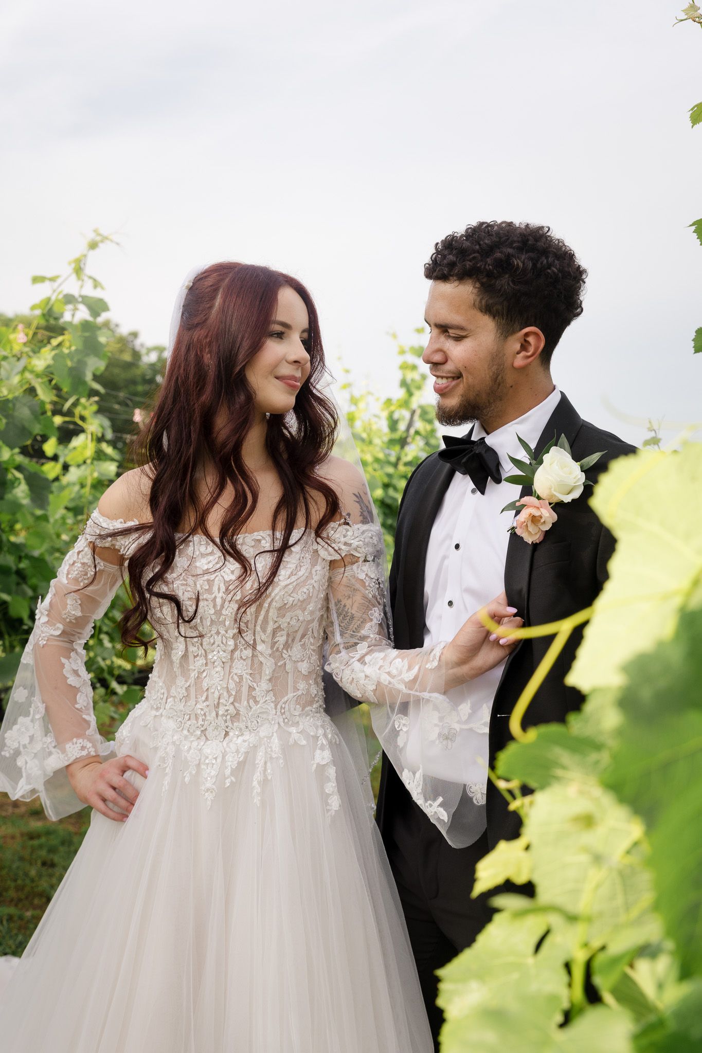 Bride in lace off-shoulder gown and groom in black tuxedo smile at each other in a lush vineyard.
