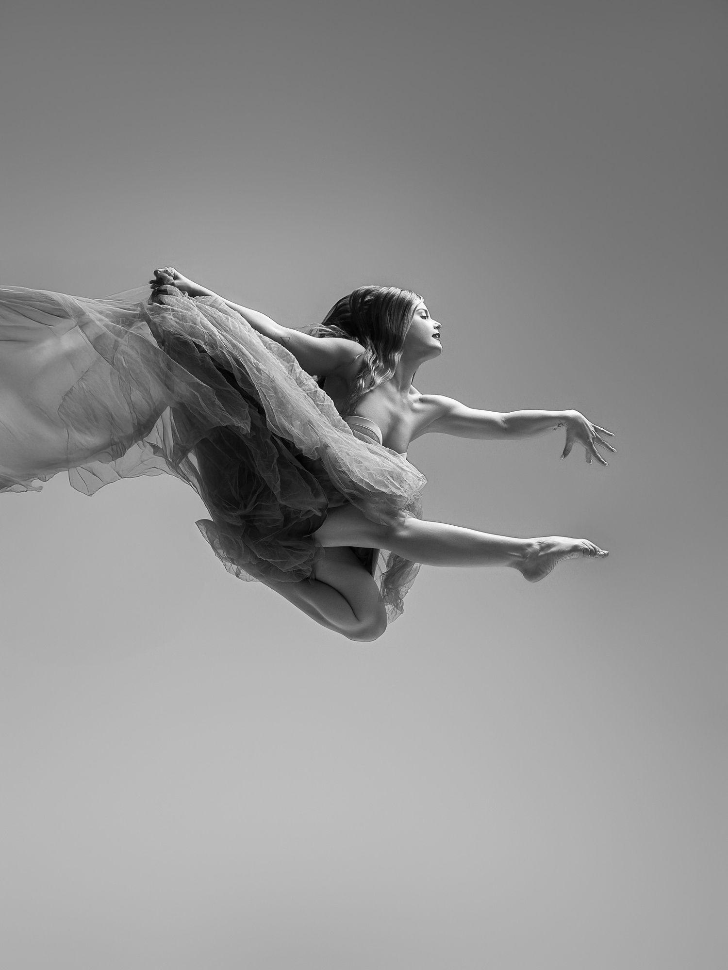 A graceful dancer in flowing white fabric leaps through the air in an elegant black and white artistic photograph.