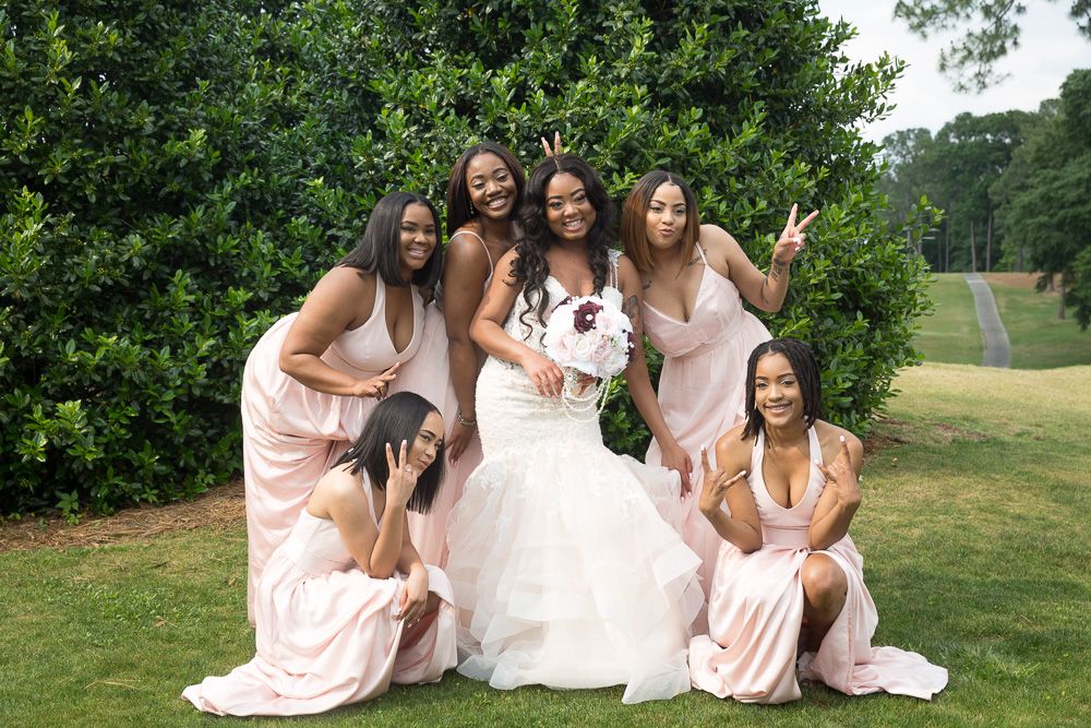 Bride in white gown poses with five bridesmaids in blush pink dresses outdoors on green lawn with lush hedges.