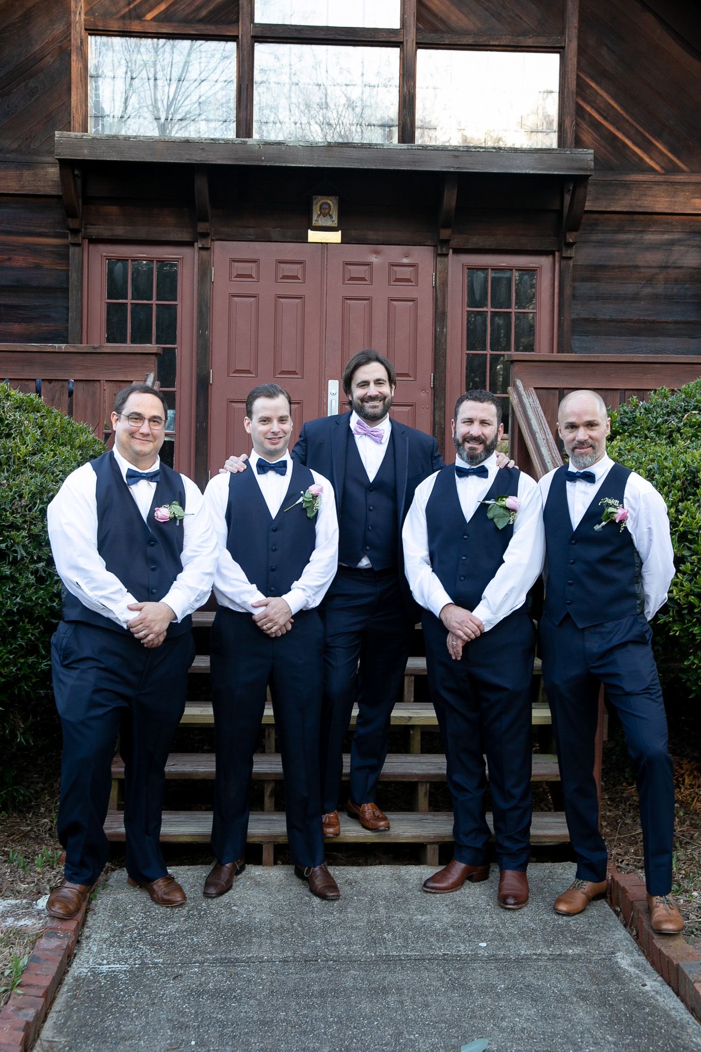 Groom and four groomsmen in navy vests and bow ties pose on church steps for wedding portrait.