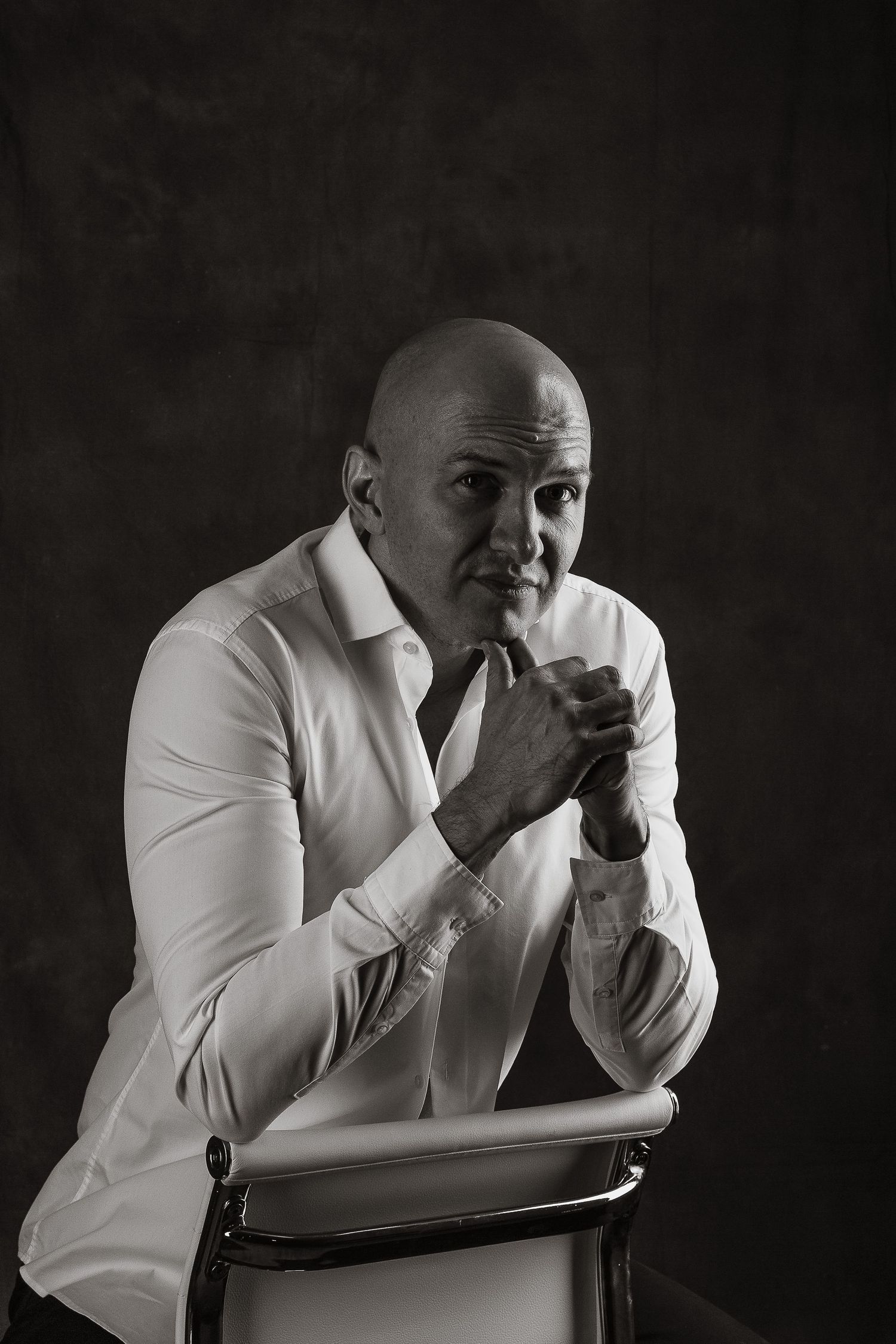 Moody black and white portrait of a man in white shirt leaning on a chair.