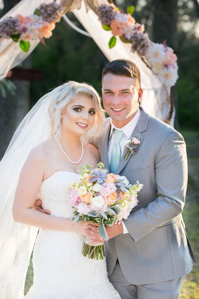 Smiling bride and groom pose under floral arch, bride holding pastel bouquet in white gown and veil, groom in gray suit.