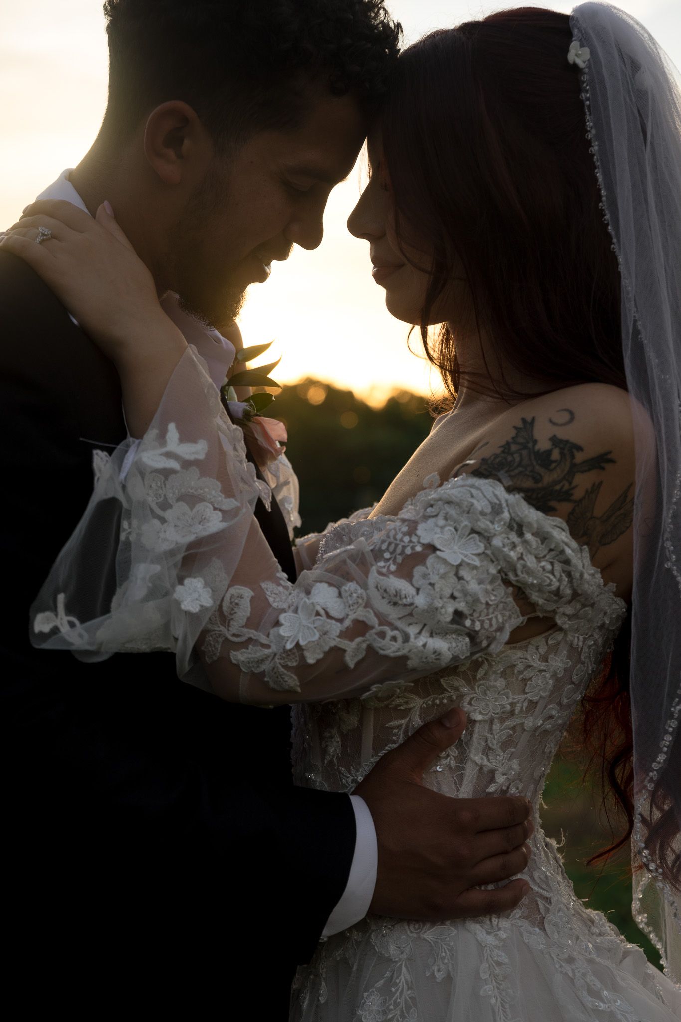 Bride and groom embracing at sunset, bride in lace wedding gown with floral details and veil, groom in black suit.