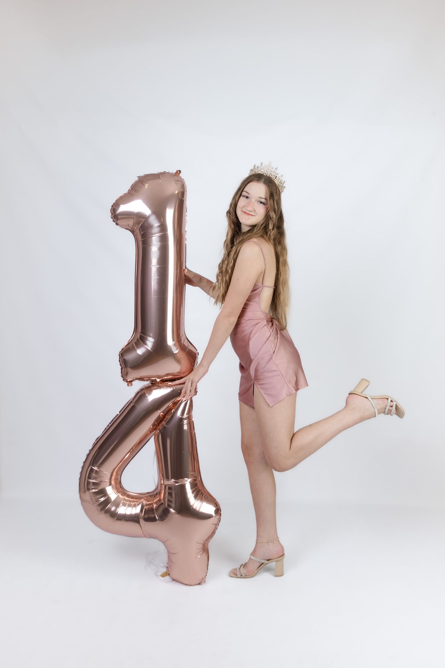 Teen girl in pink romper and tiara poses with rose gold number 14 balloons celebrating her birthday.