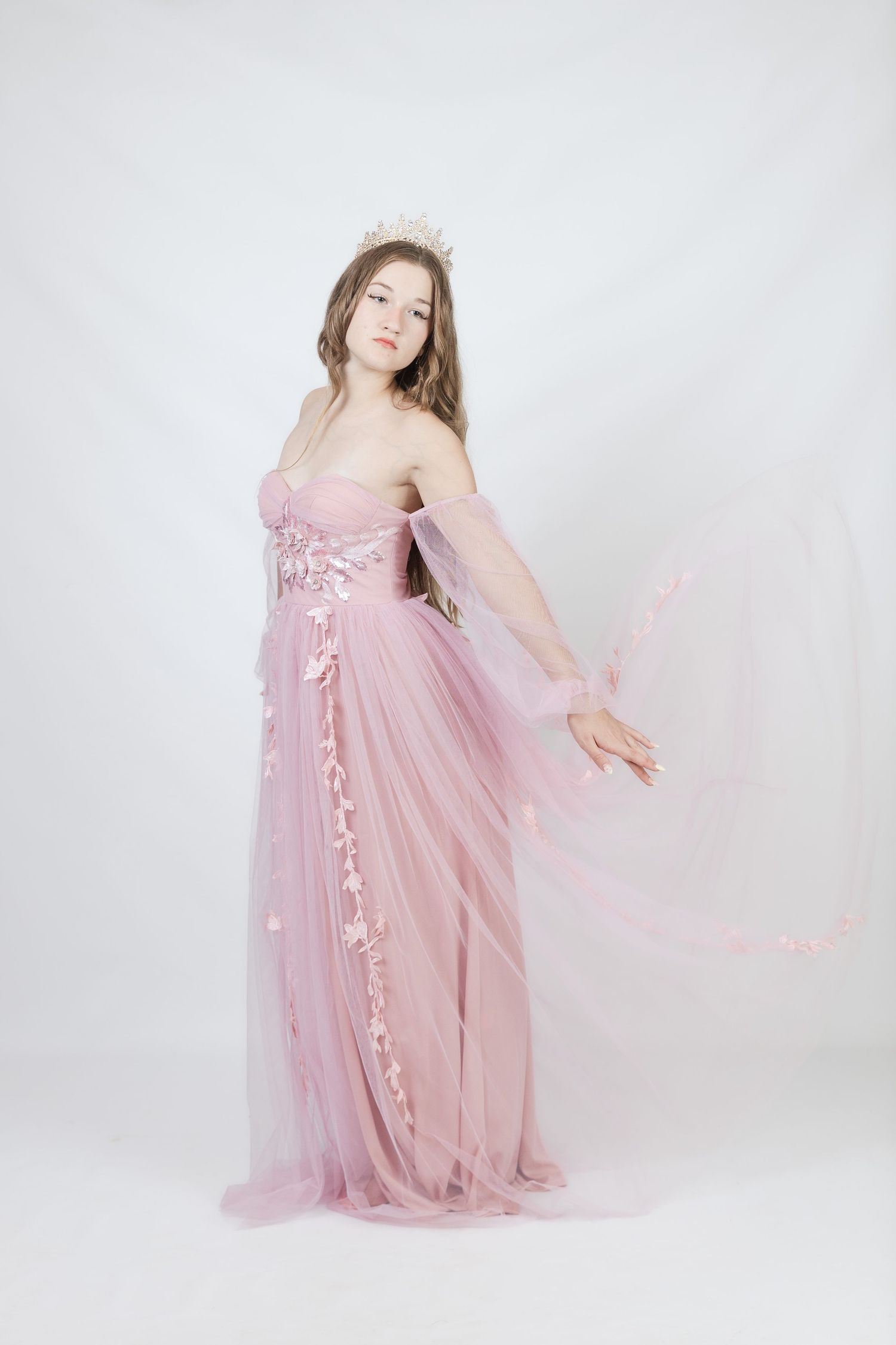 Young woman in a flowing pink tulle gown with floral details and a tiara poses gracefully on white background.