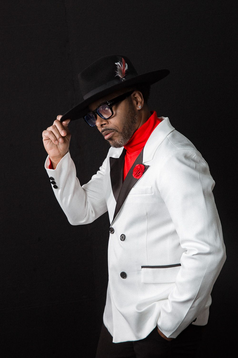 Stylish figure in white blazer and black hat poses dramatically against dark background.