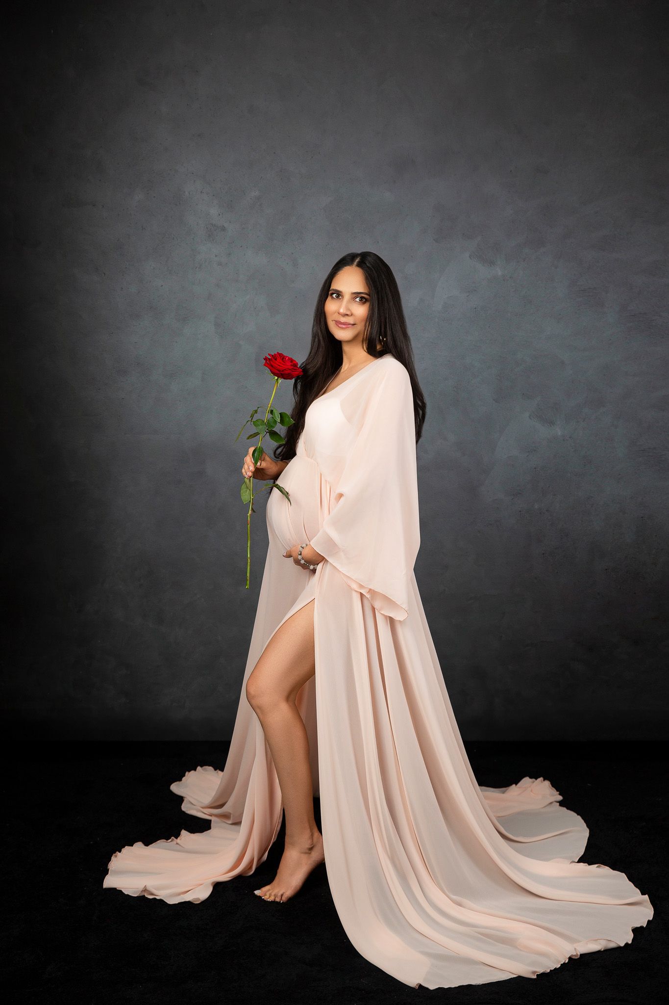 A flowing blush pink maternity gown drapes elegantly against a dark studio backdrop.