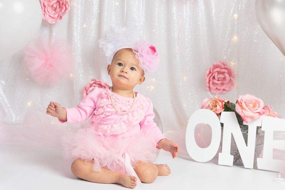 Baby girl in pink tutu dress sits beside ONE sign with floral decorations for first birthday cake smash photo.