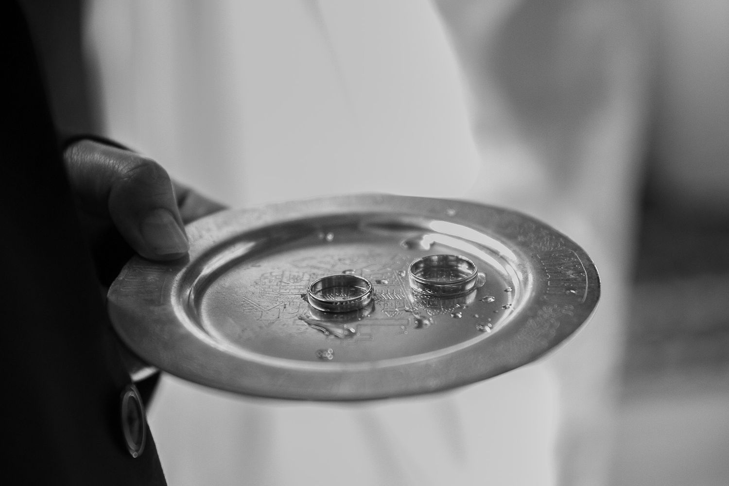 Black and white photo of two wedding rings on a silver plate held by a person during a wedding ceremony.