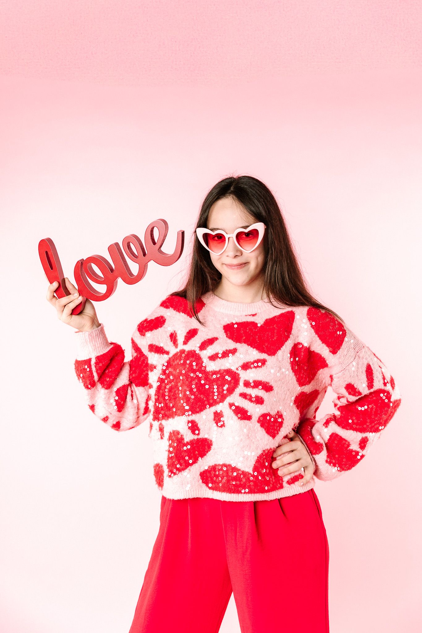 Heart-patterned sweater styled with red pants and love prop against pink backdrop.