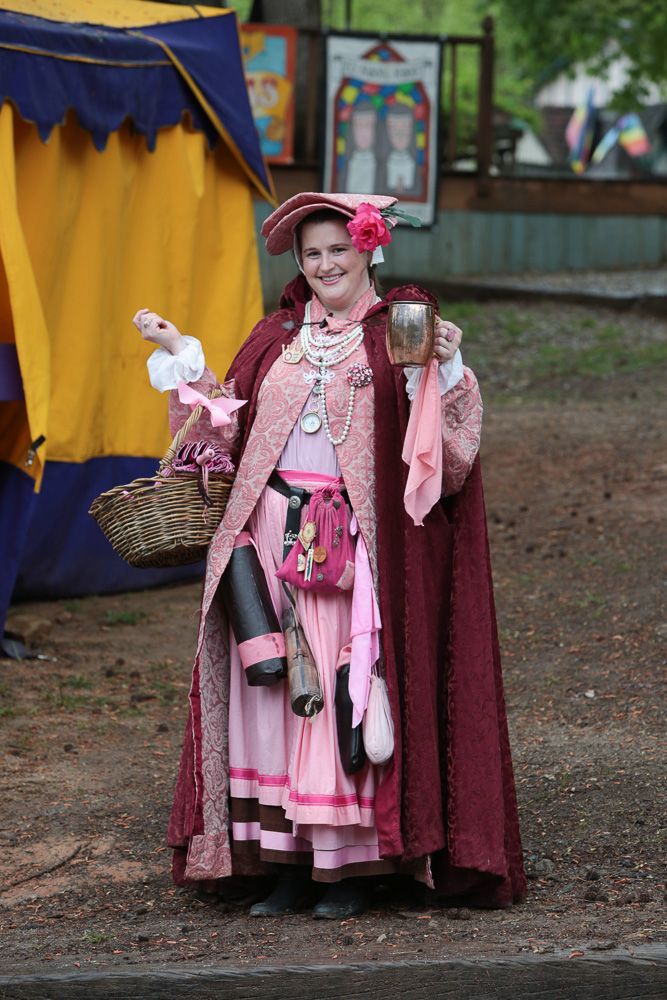 Renaissance fair performers in period costumes with pink and burgundy dresses walking through a festival ground.