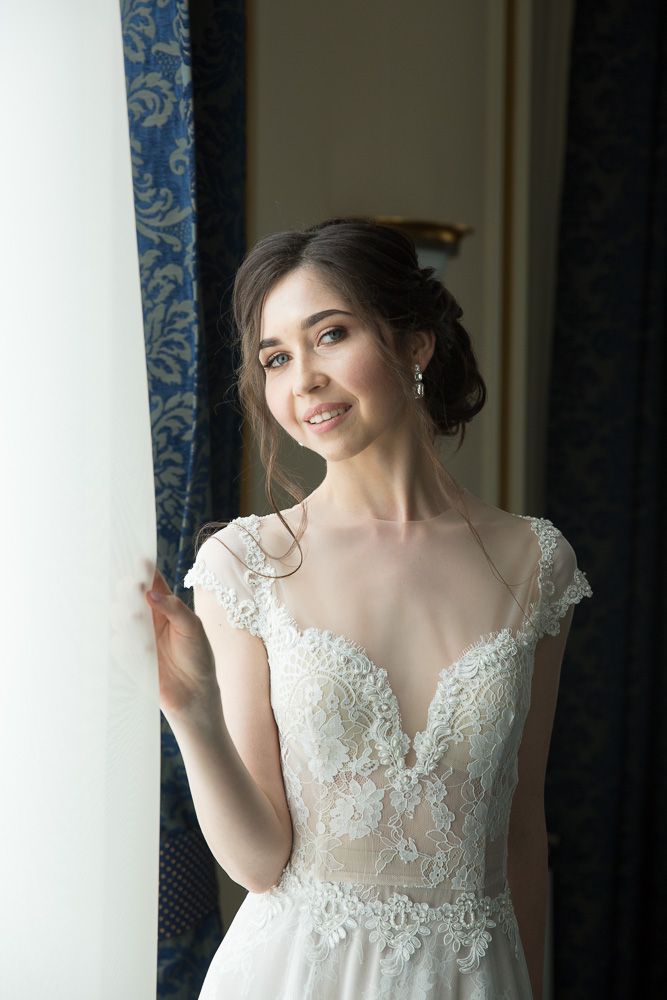 Beautiful smiling bride in lace wedding dress standing by a window with blue curtains.
