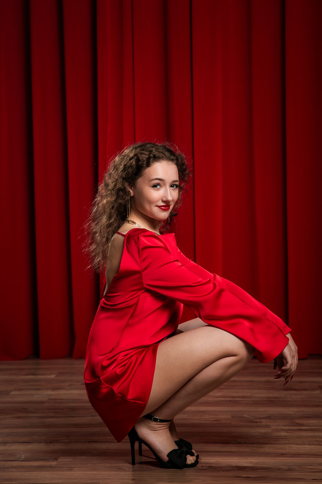 Performer in red outfit strikes dramatic crouching poses on wooden floor with red curtain background.