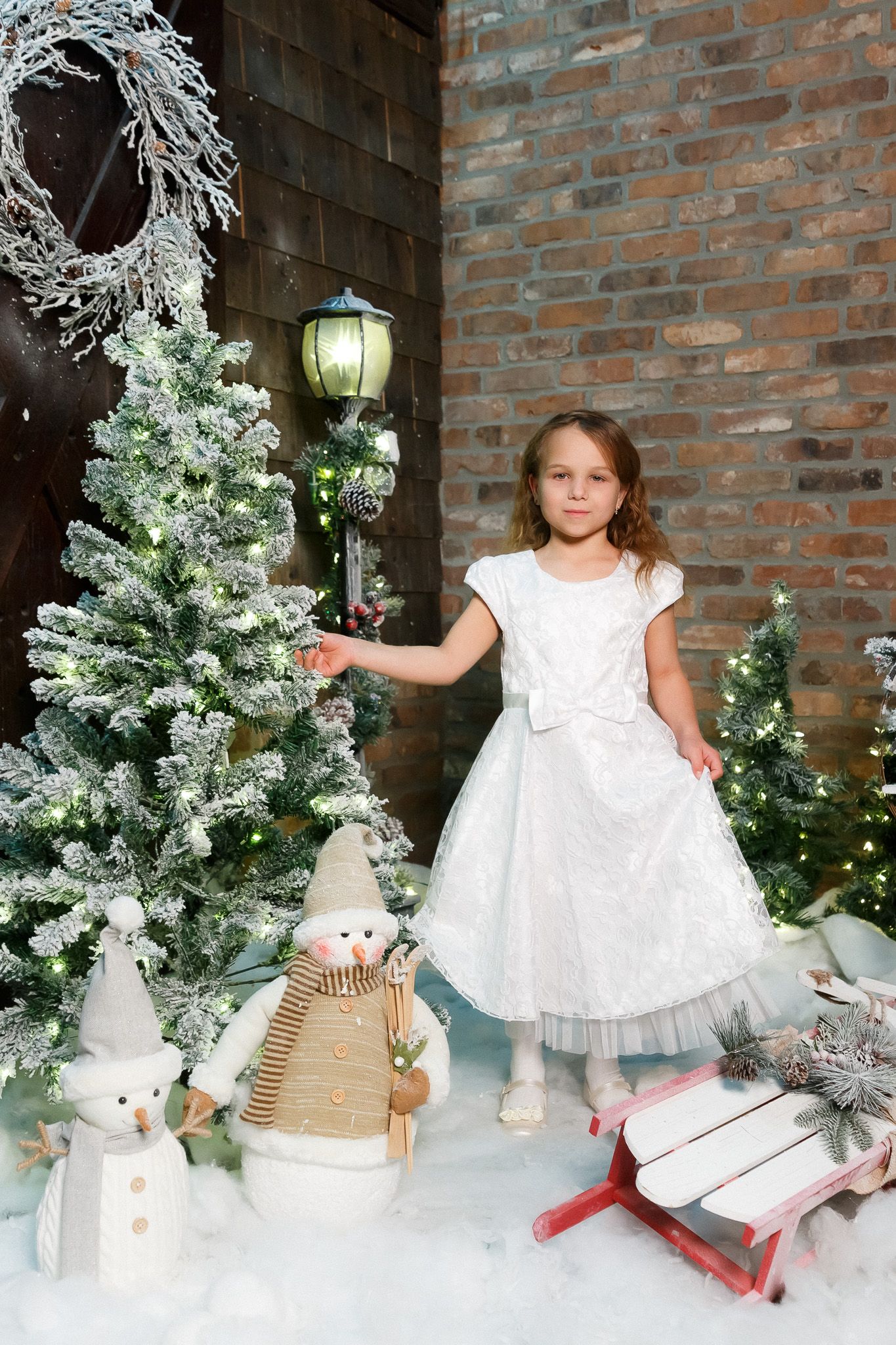 Winter wonderland scene with decorated Christmas trees, snowman decor, and white dress against brick wall.