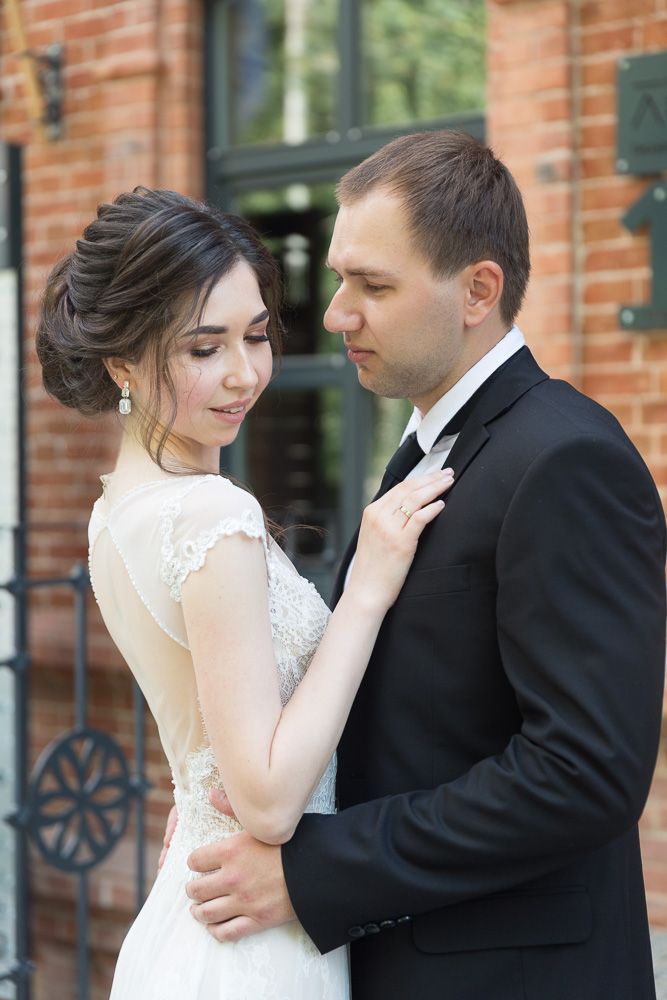 Bride and groom embracing outdoors near brick building, bride in lace dress with elegant updo, groom in black suit.