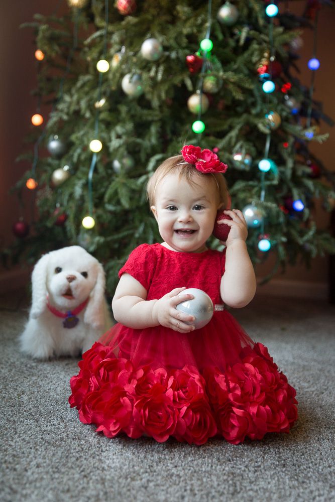 Baby in red ruffled dress sits on carpet near decorated Christmas tree with white plush toy.
