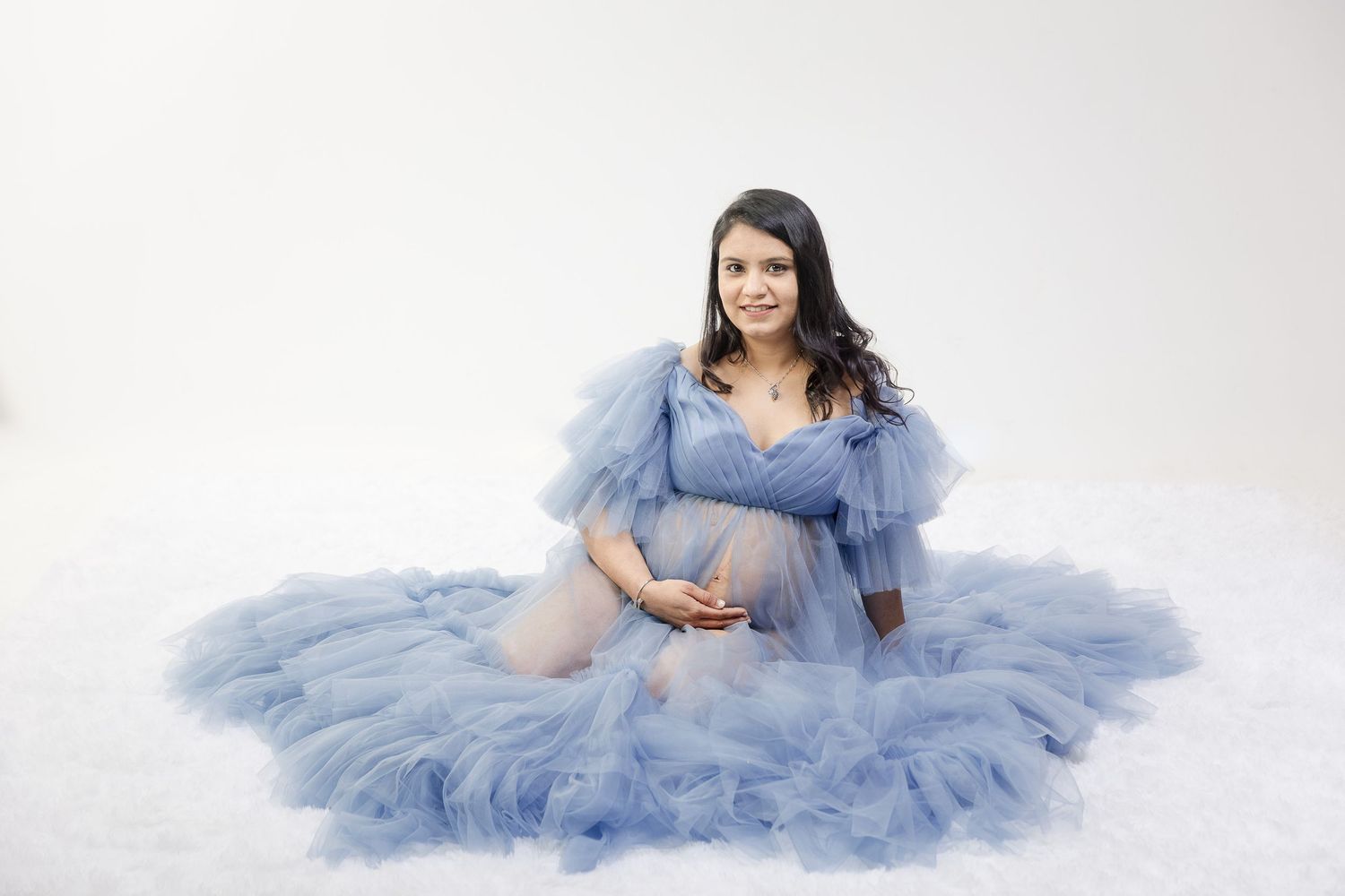 Pregnant woman in a flowing blue tulle gown sits gracefully on a white studio floor for a maternity photo shoot.