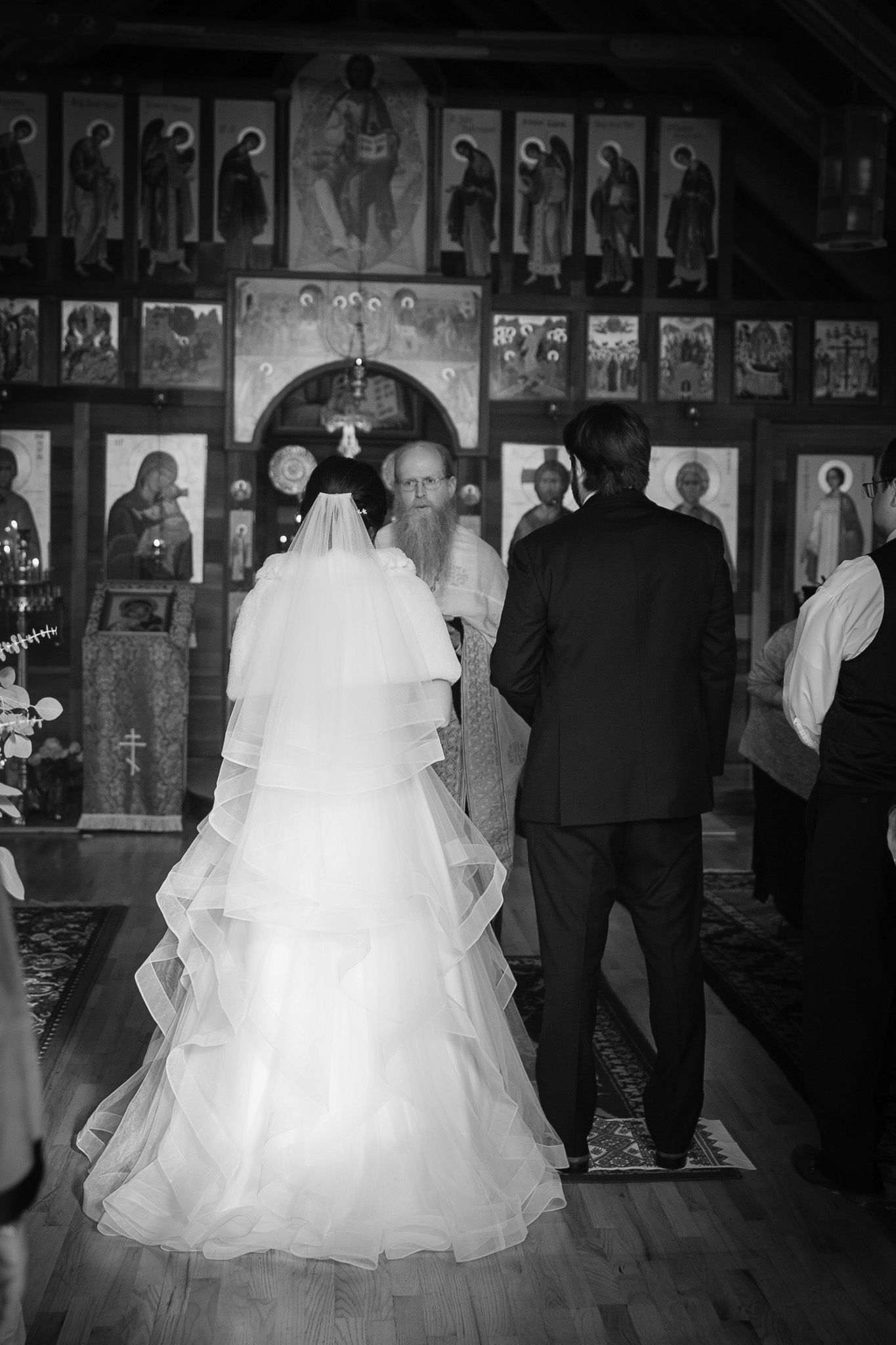 Black and white photo of a bride and groom standing before an Orthodox priest at a church wedding ceremony.