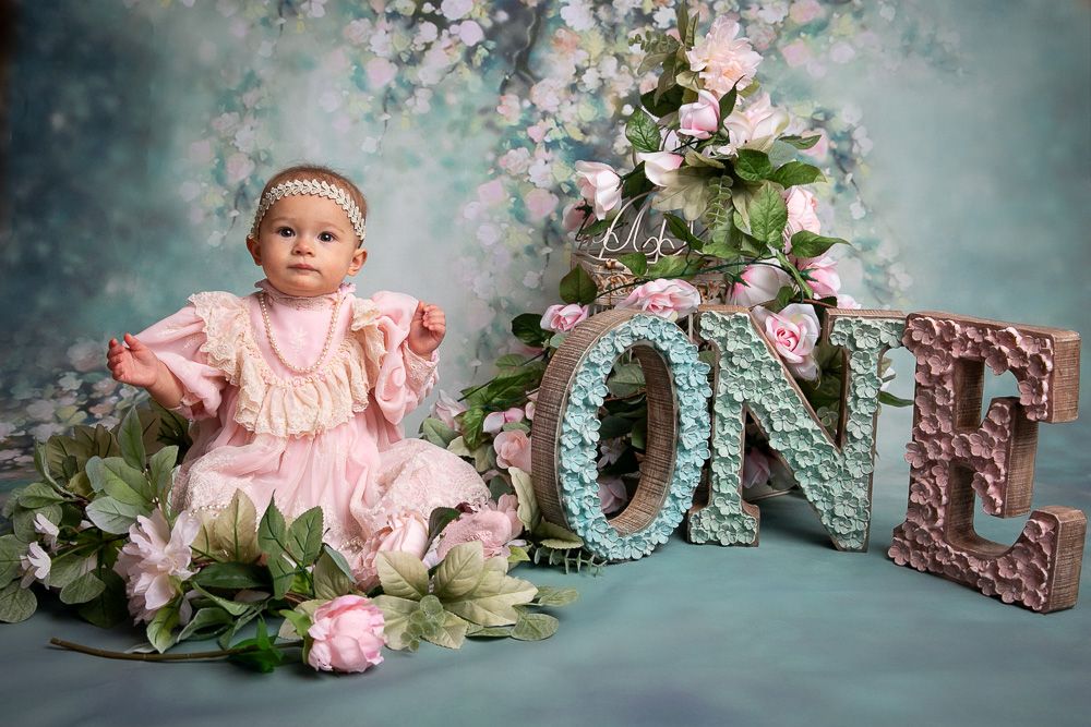Baby girl in pink dress sits beside decorative ONE letters with flowers for her first birthday photo session.